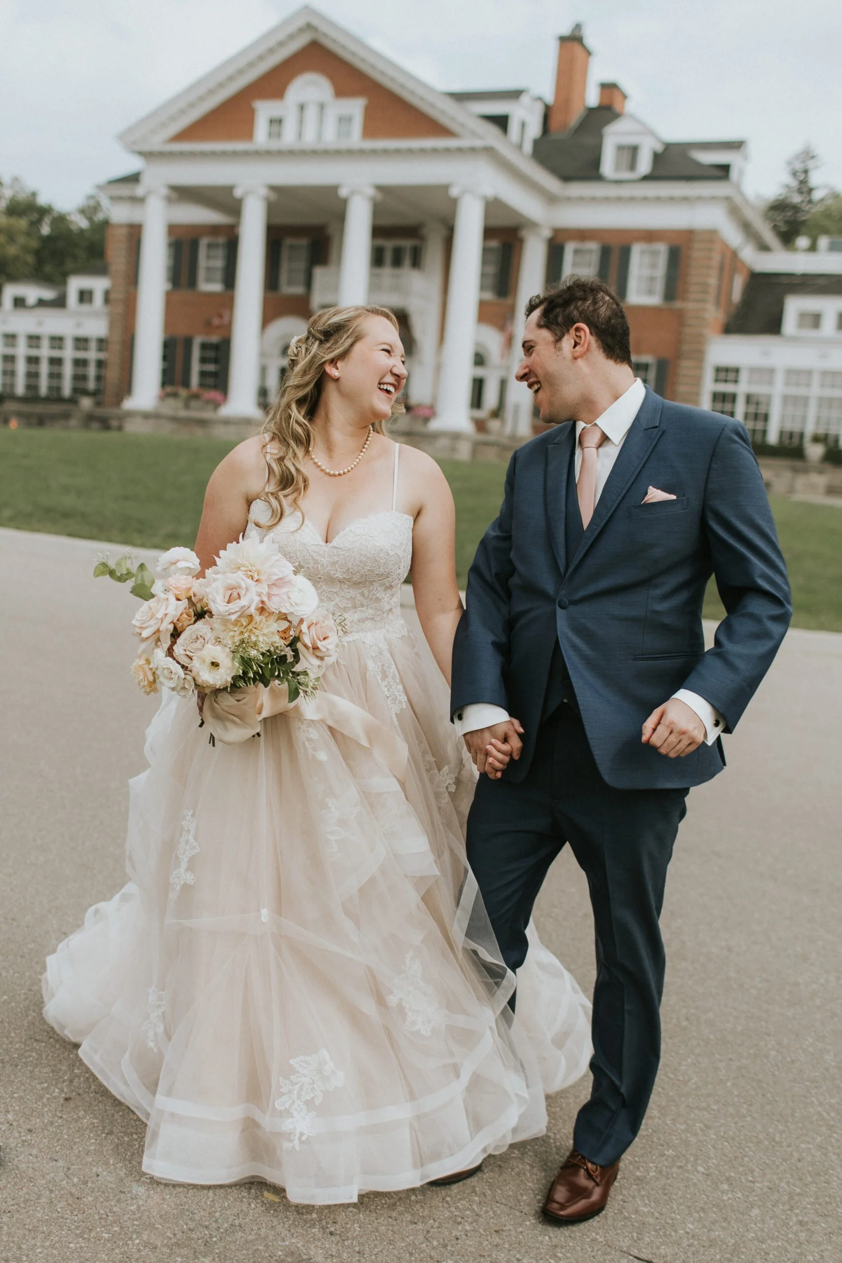 A bride and groom walking hand in hand outdoors in front of a large mansion, smiling and looking at each other. The bride wears a white lace wedding dress and holds a bouquet, while the groom is dressed in a navy blue suit with a tie.