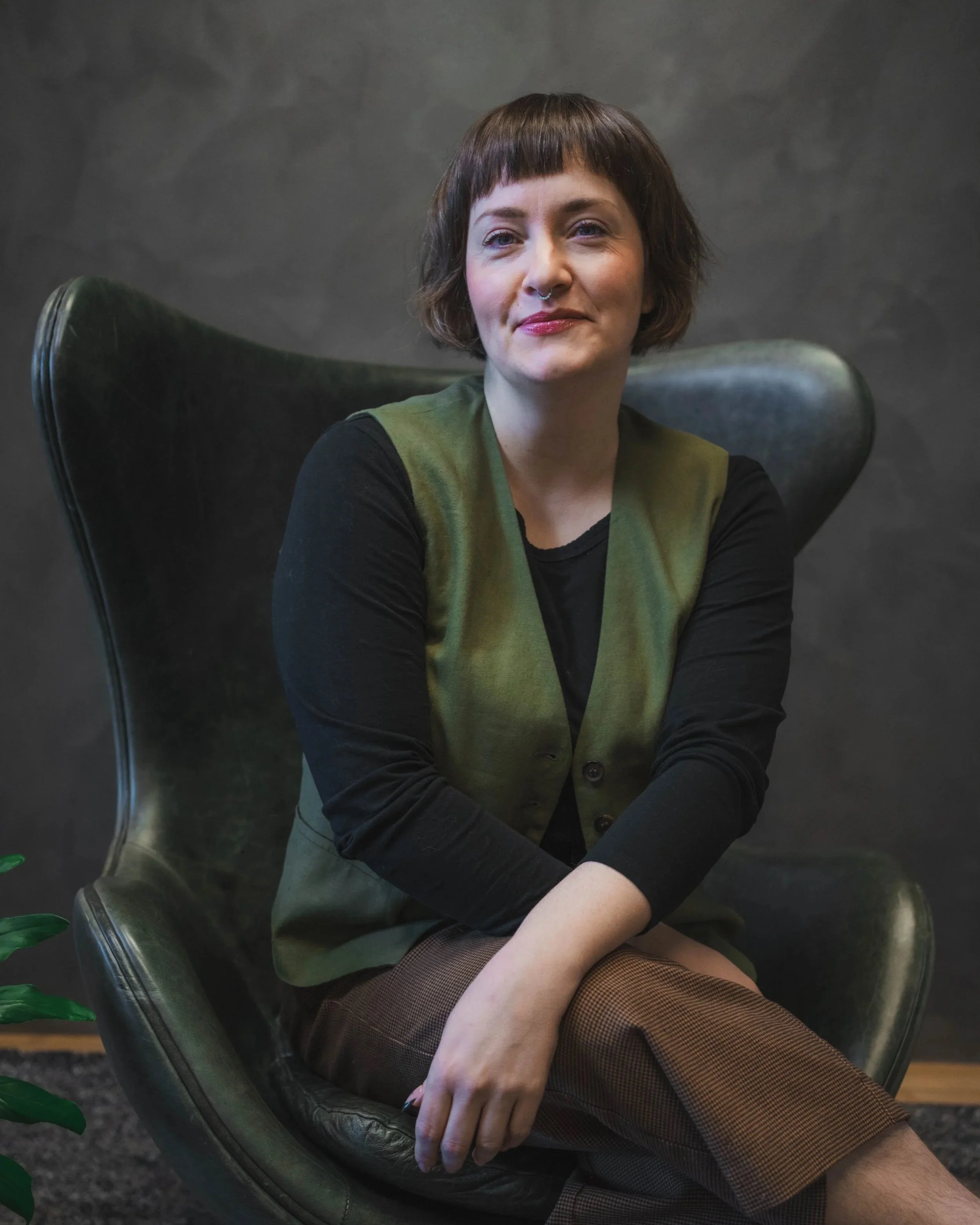 A young woman with short brown hair and a septum piercing, sitting in a modern green leather chair, wearing a black long-sleeve shirt and green vest.