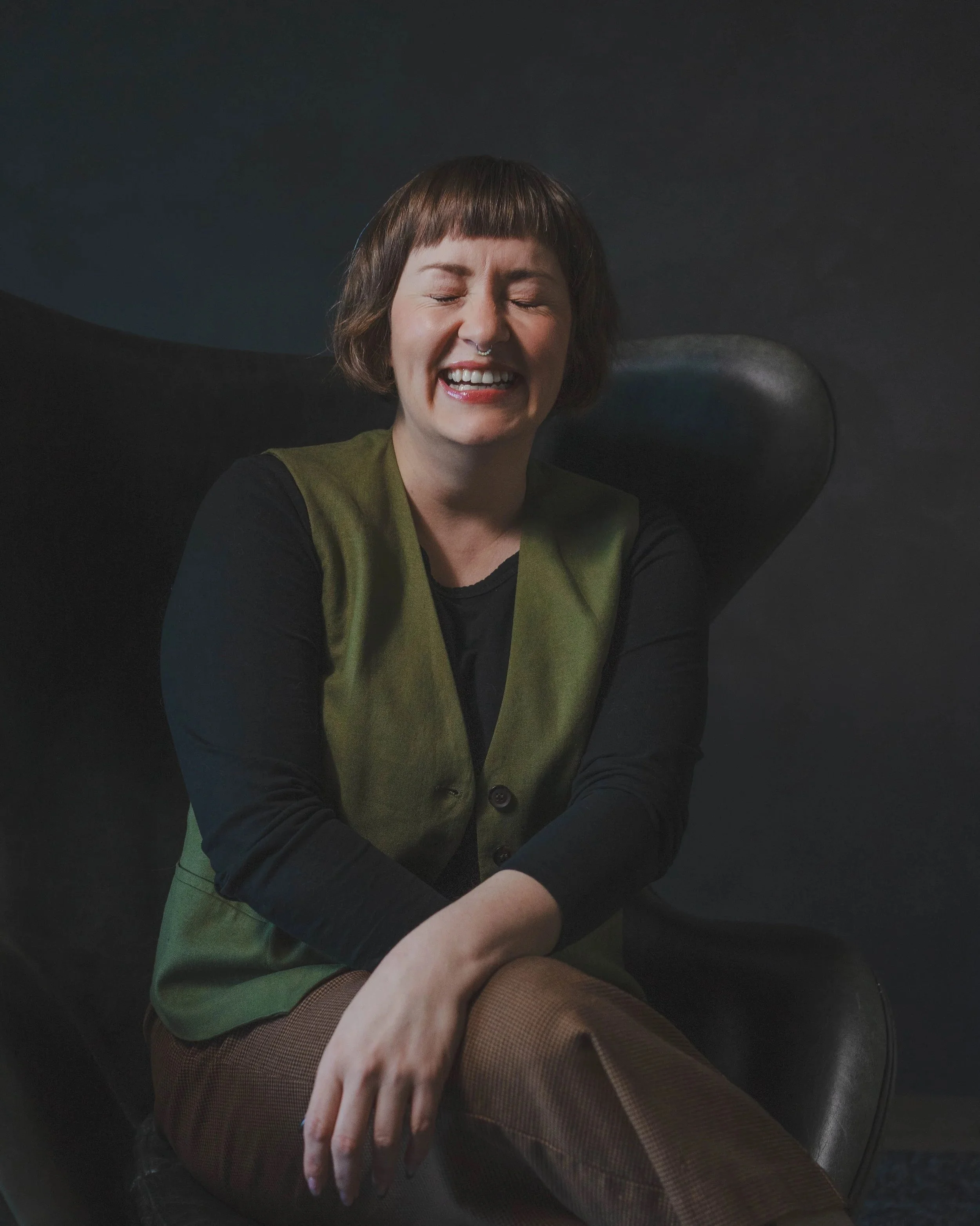A woman with short brown hair and a septum piercing sitting on a black chair, smiling with her eyes closed, wearing a green vest over a black long-sleeve shirt.