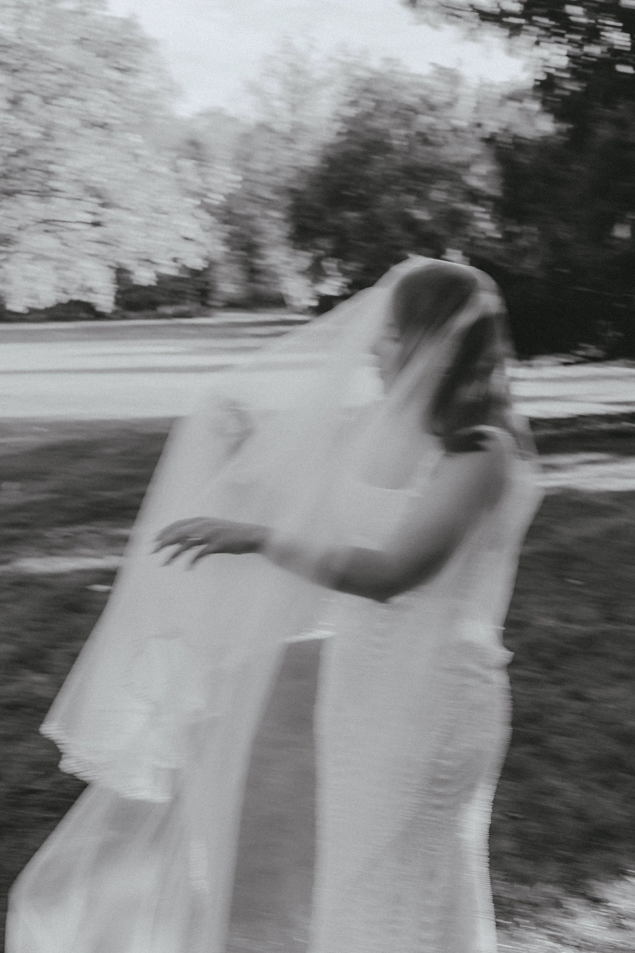 A blurry black and white photograph of a woman standing outdoors near trees.