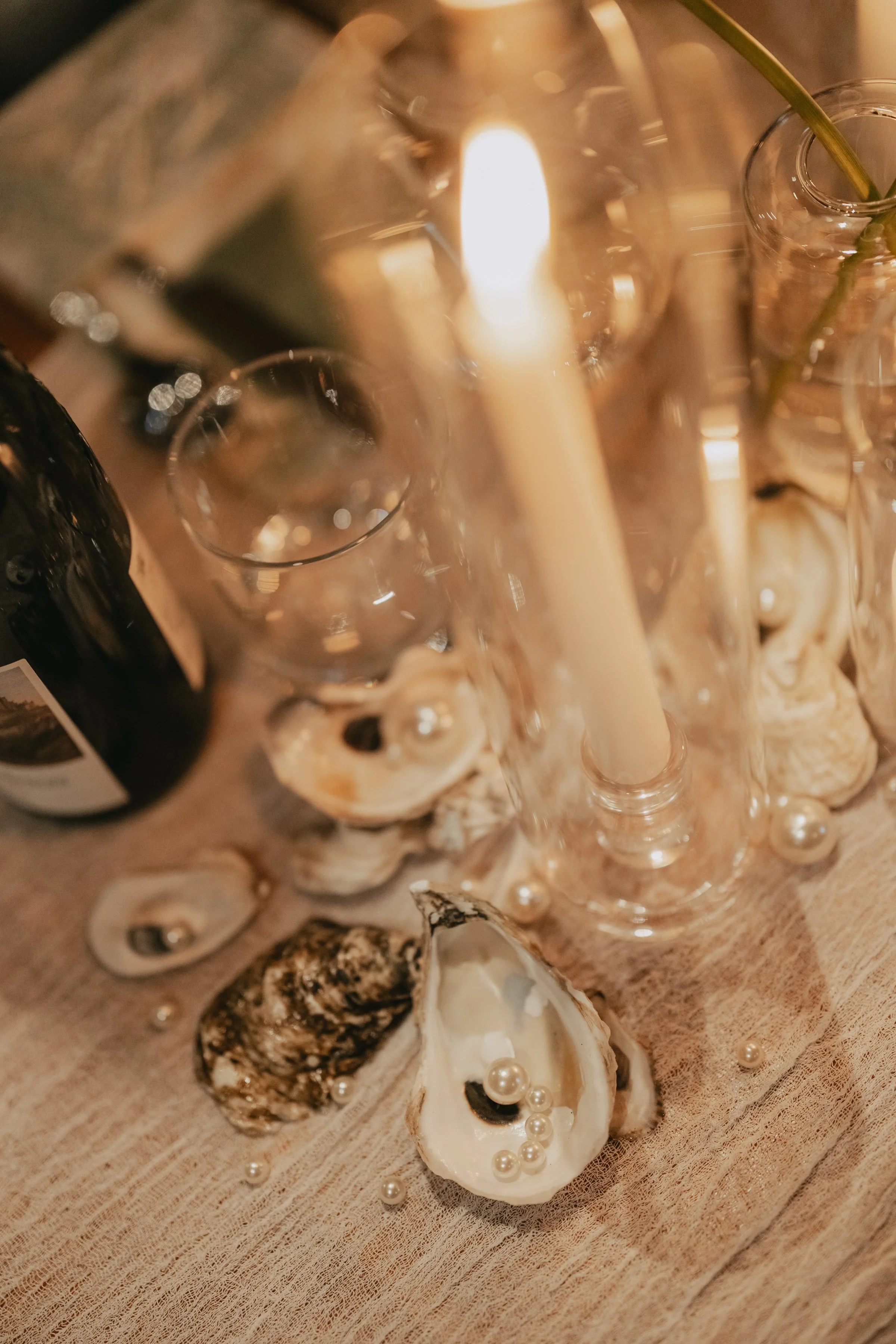 Close-up of a candlelit table with seashells, a glass bottle with green stems, a glass, and a bottle, creating an elegant, coastal-themed setting.