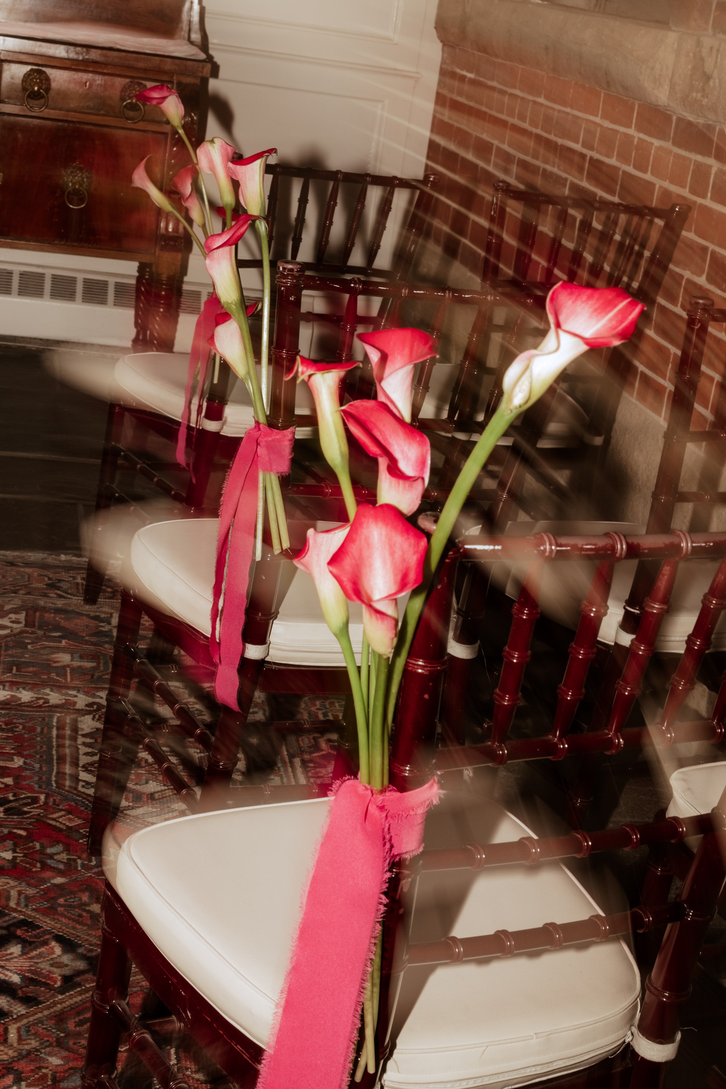 A bouquet of pink and white calla lilies tied with a pink ribbon is placed on a wooden chair with a white cushion, in a room with a patterned rug, brick wall, and wooden furniture.