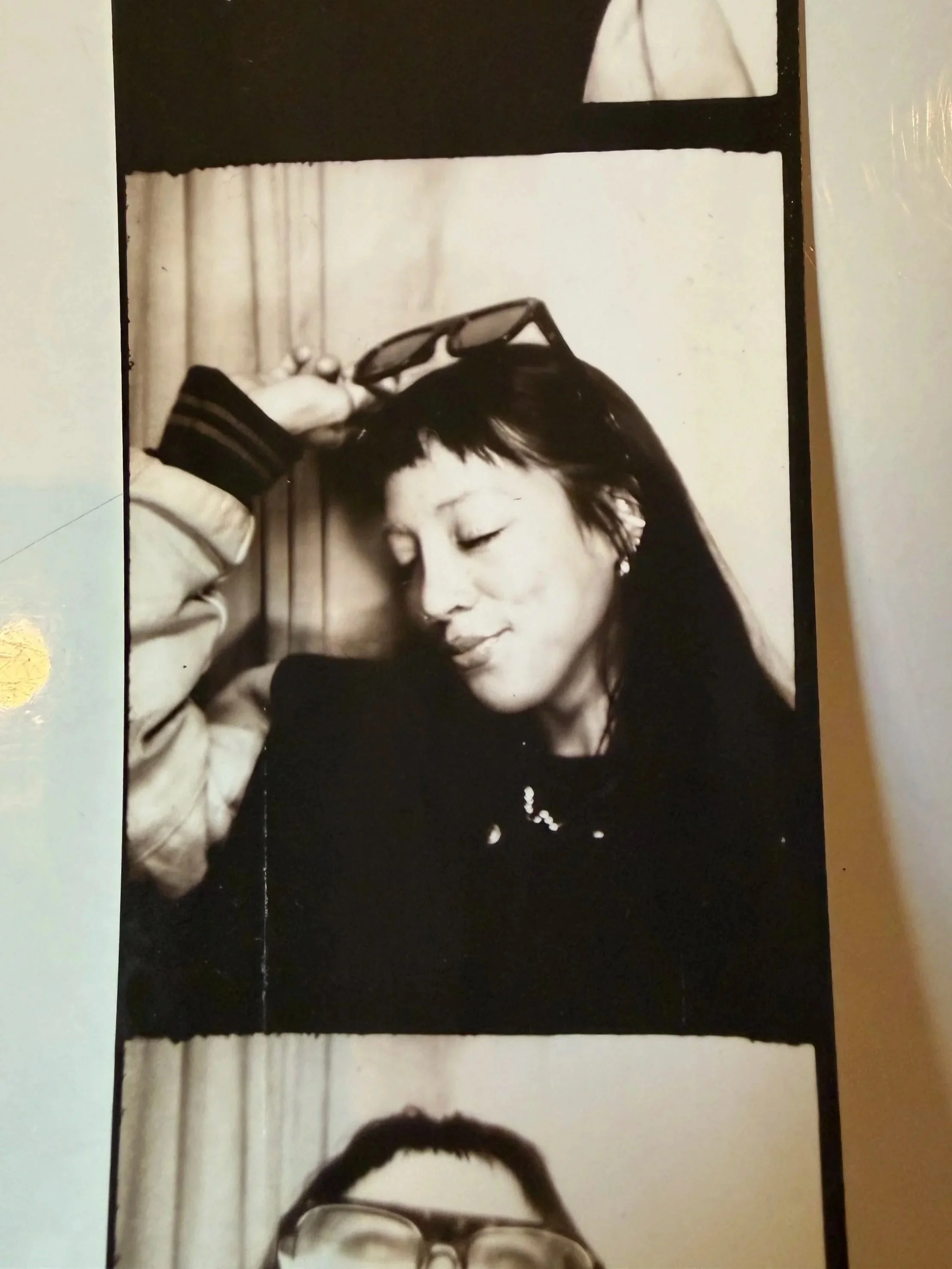 Black and white photo of a woman with closed eyes, holding sunglasses on her head, with a slight smile, sitting against a curtain background.