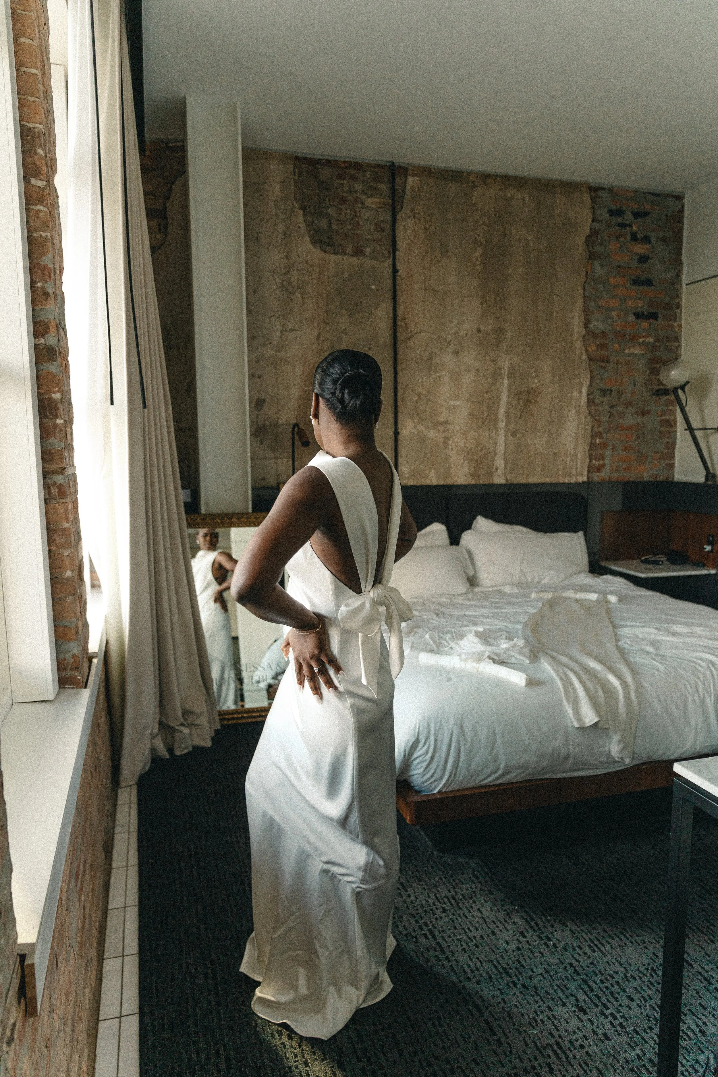 A woman in an elegant white dress standing in a hotel room, looking out the window, with a bed and a large mirror in the background. Hotel Haya, Ybor City Fl. Luxury wedding, luxury brides, Nyc editorial