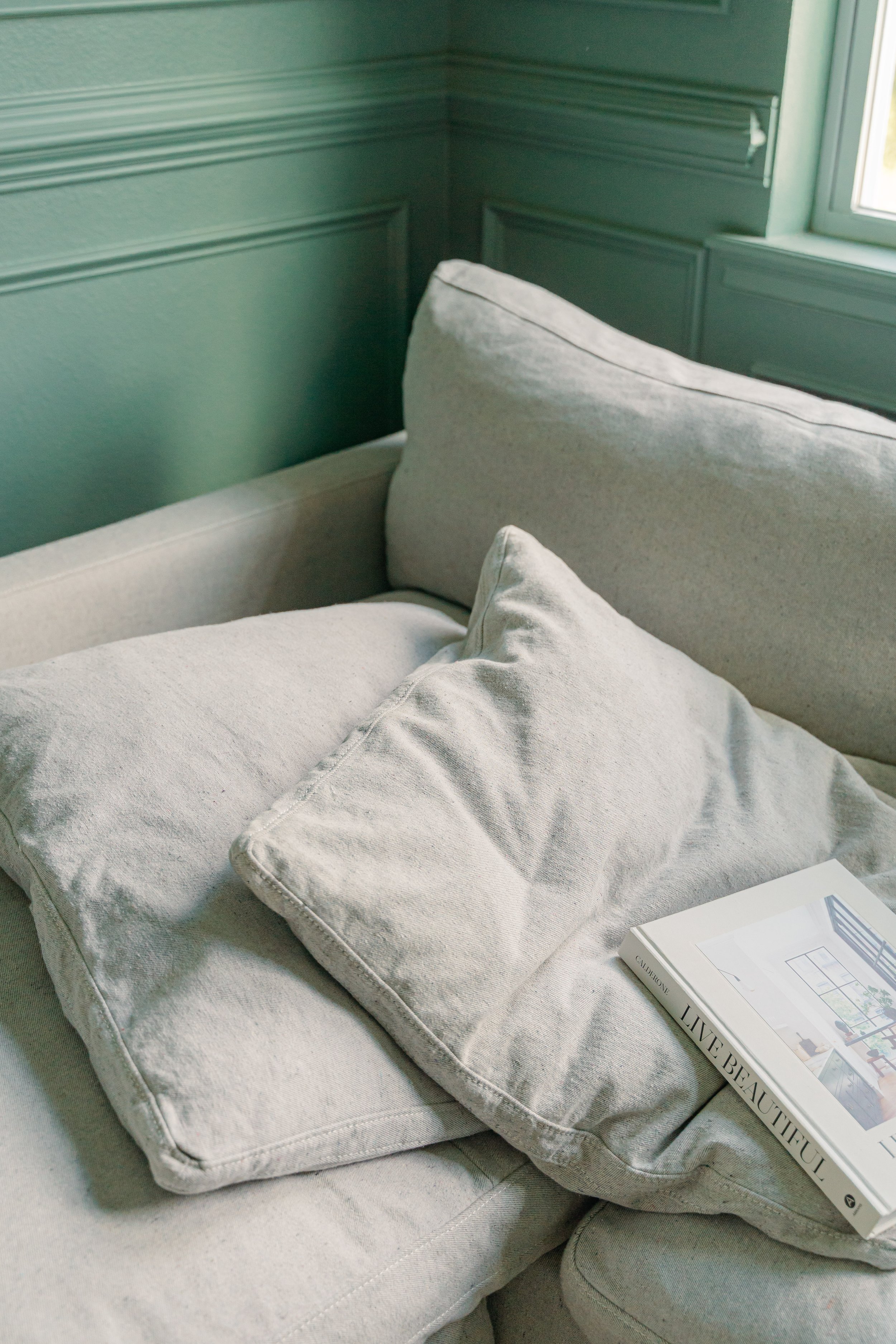 A cream-colored sofa with light-colored pillows, placed next to a window with emerald green walls and decorative molding.
