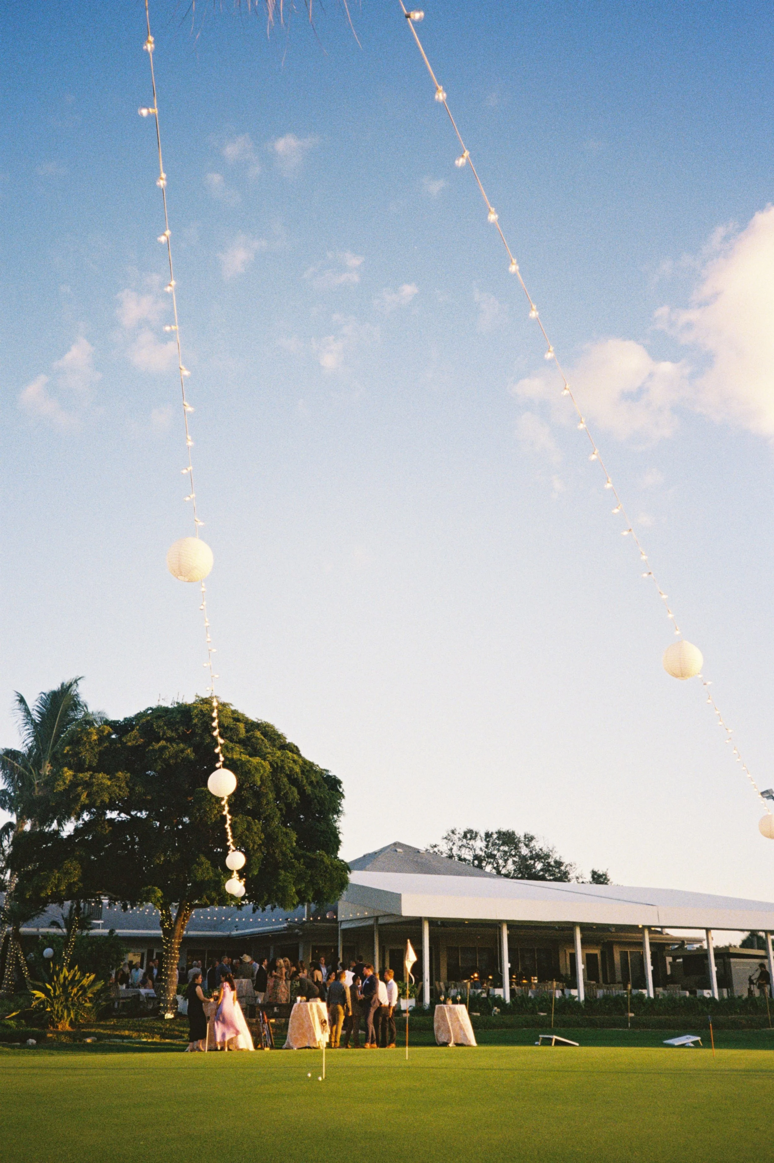 People gathered at a social event on a golf course, near a clubhouse, with string lights and paper lanterns hanging overhead during the evening.