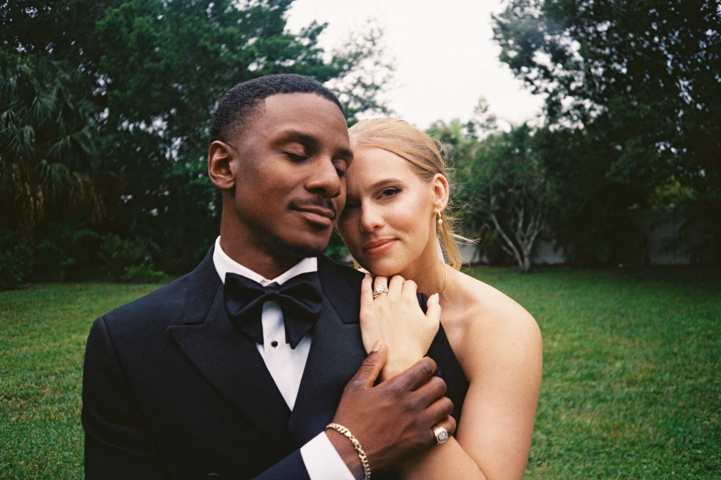 A close-up of a couple dressed in formal wedding attire, embracing outdoors on a grassy area with trees in the background.