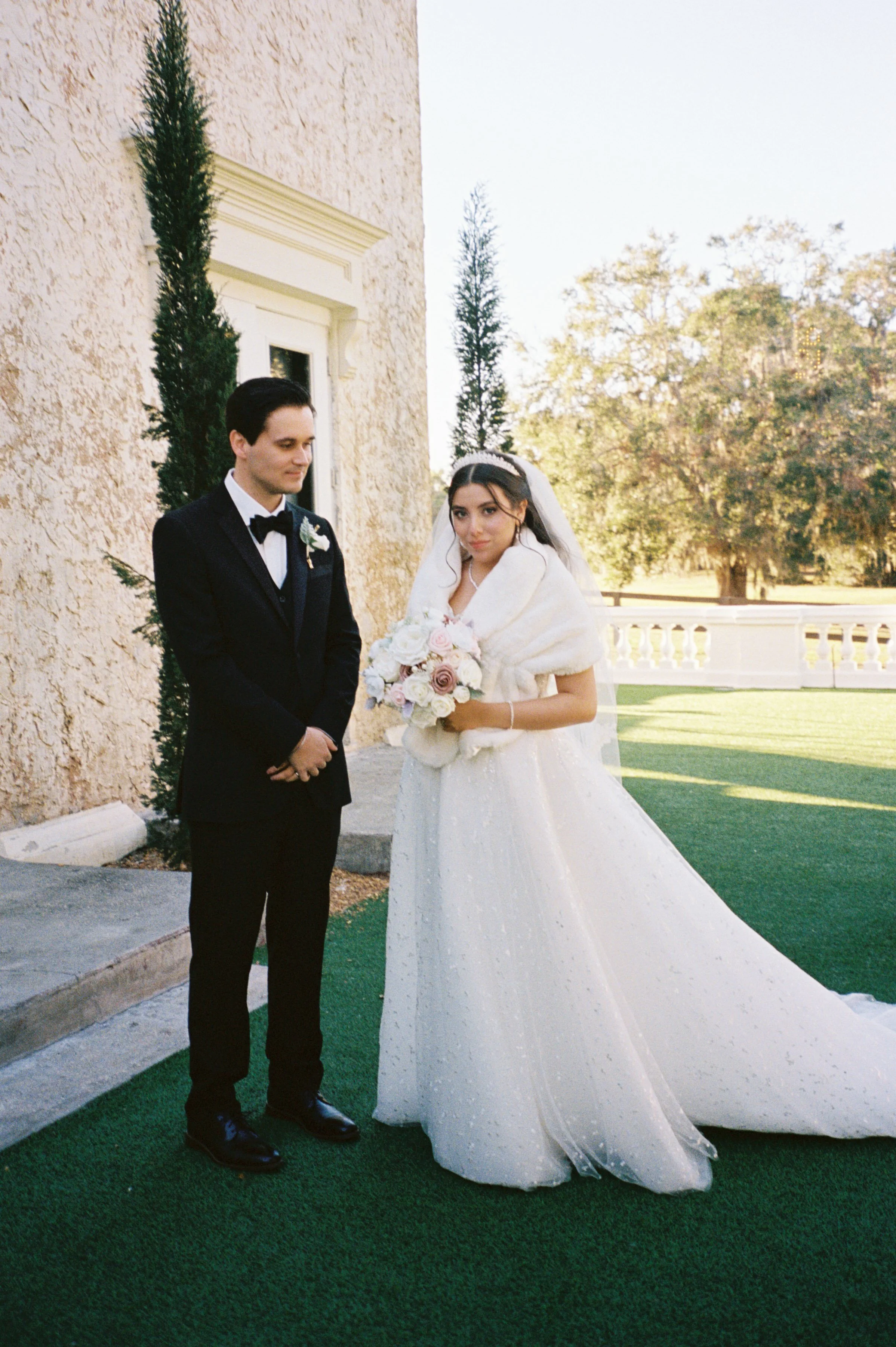 A bride and groom standing outdoors in wedding attire, with the bride holding a bouquet of roses, near a textured beige wall and some tall greenery. 35mm film photos. Bella Cosa wedding venue. Classic Bride style, Winter Wedding. NY love, FL lovers