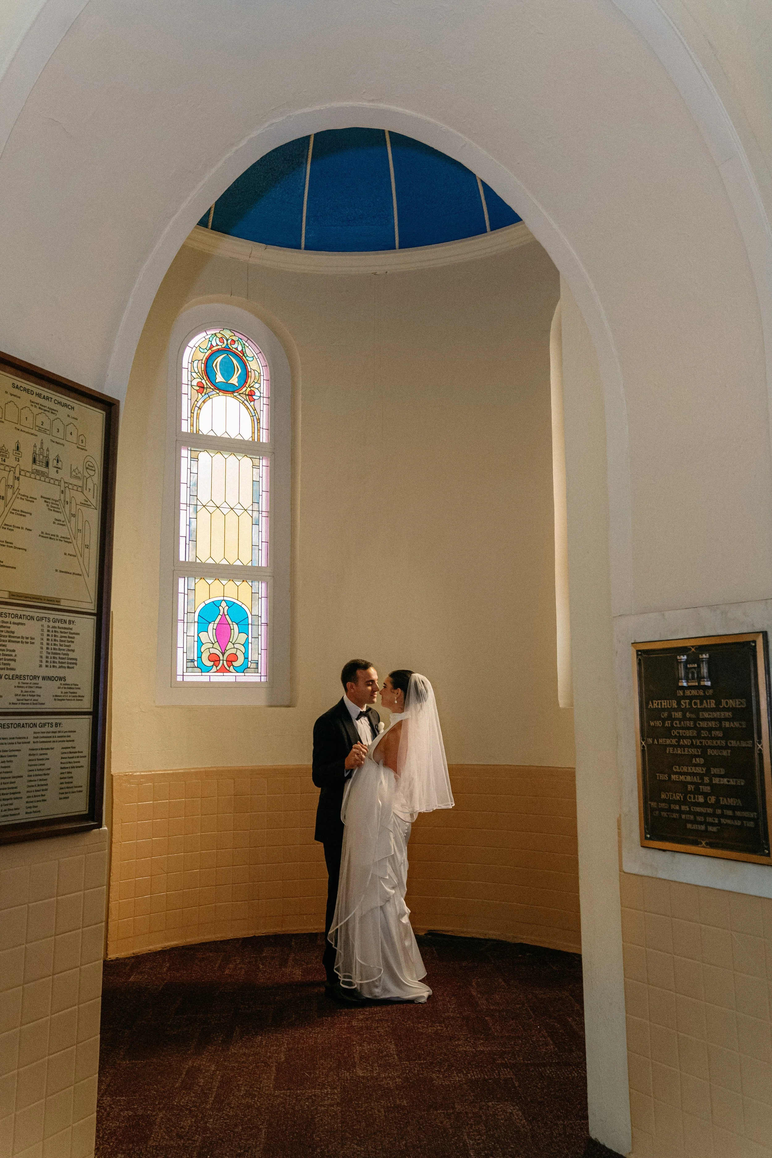 Wedding couple inside a church, standing close and looking at each other, with stained glass windows behind them.