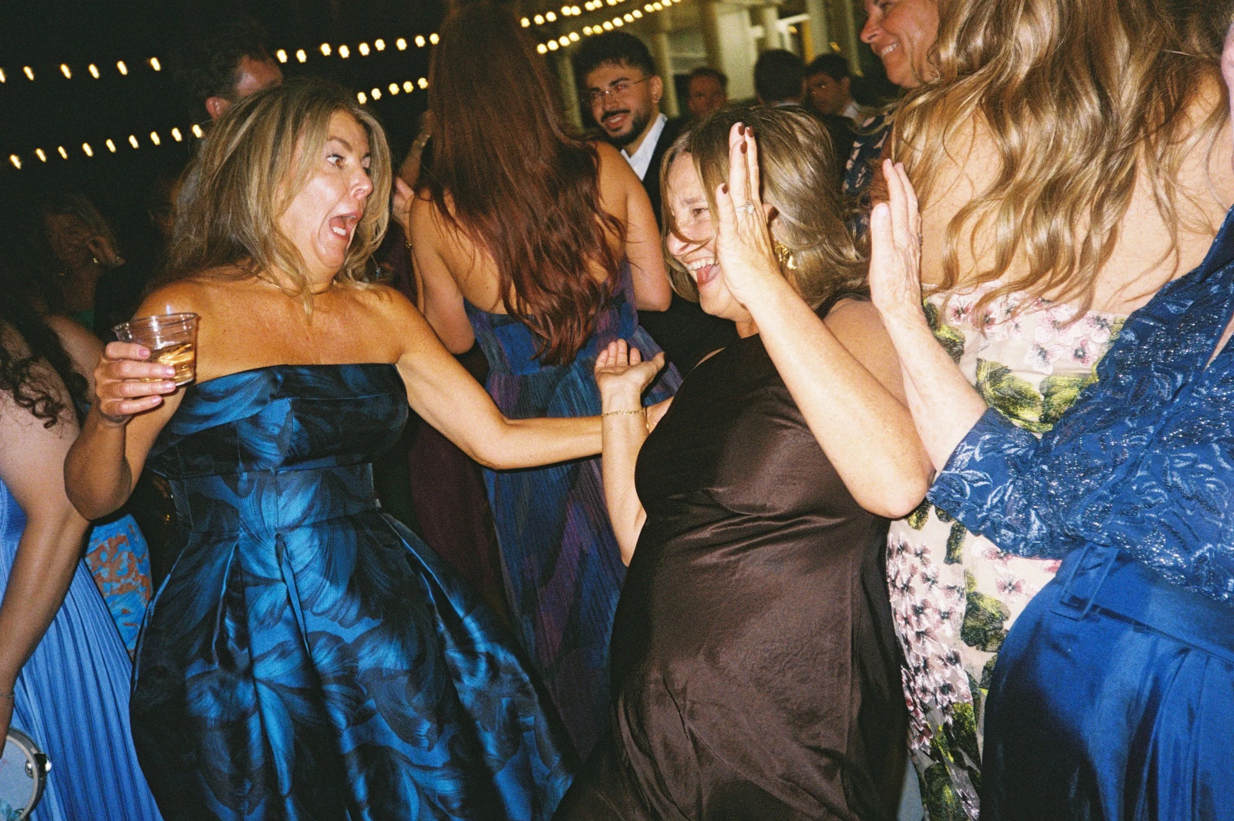 Group of women dancing and celebrating at a party or wedding reception, with string lights in the background.