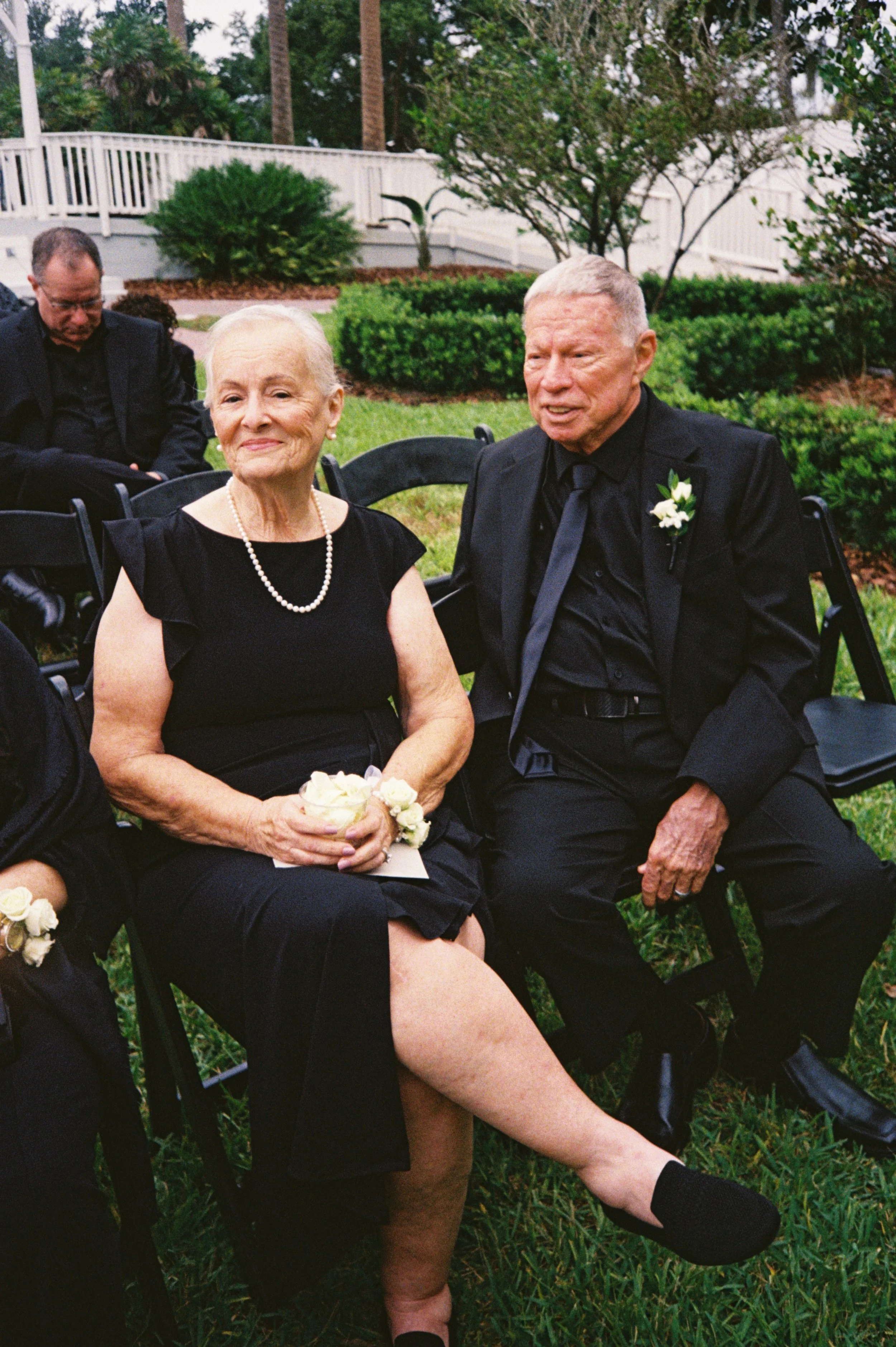 An elderly woman and man, dressed in black formal attire, sitting outdoors at a wedding or formal event. The woman is wearing a black dress with ruffled sleeves, a pearl necklace, and holding a small bouquet, with one leg crossed over the other. The man is in a black suit with a boutonnière, sitting next to her on a black folding chair.