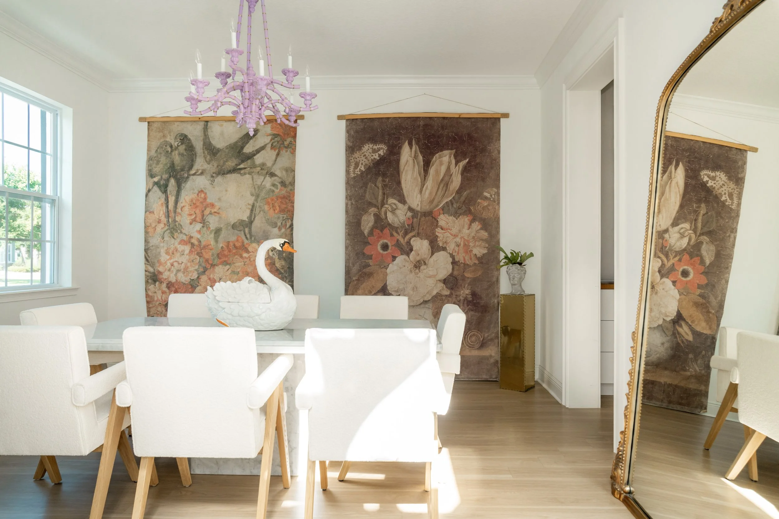 A bathroom with black and white leafy wallpaper, white tiled walls, a white pedestal sink with gold fixtures, a mirror, a plant, a wooden stool with a white container and a book, and a white toilet.