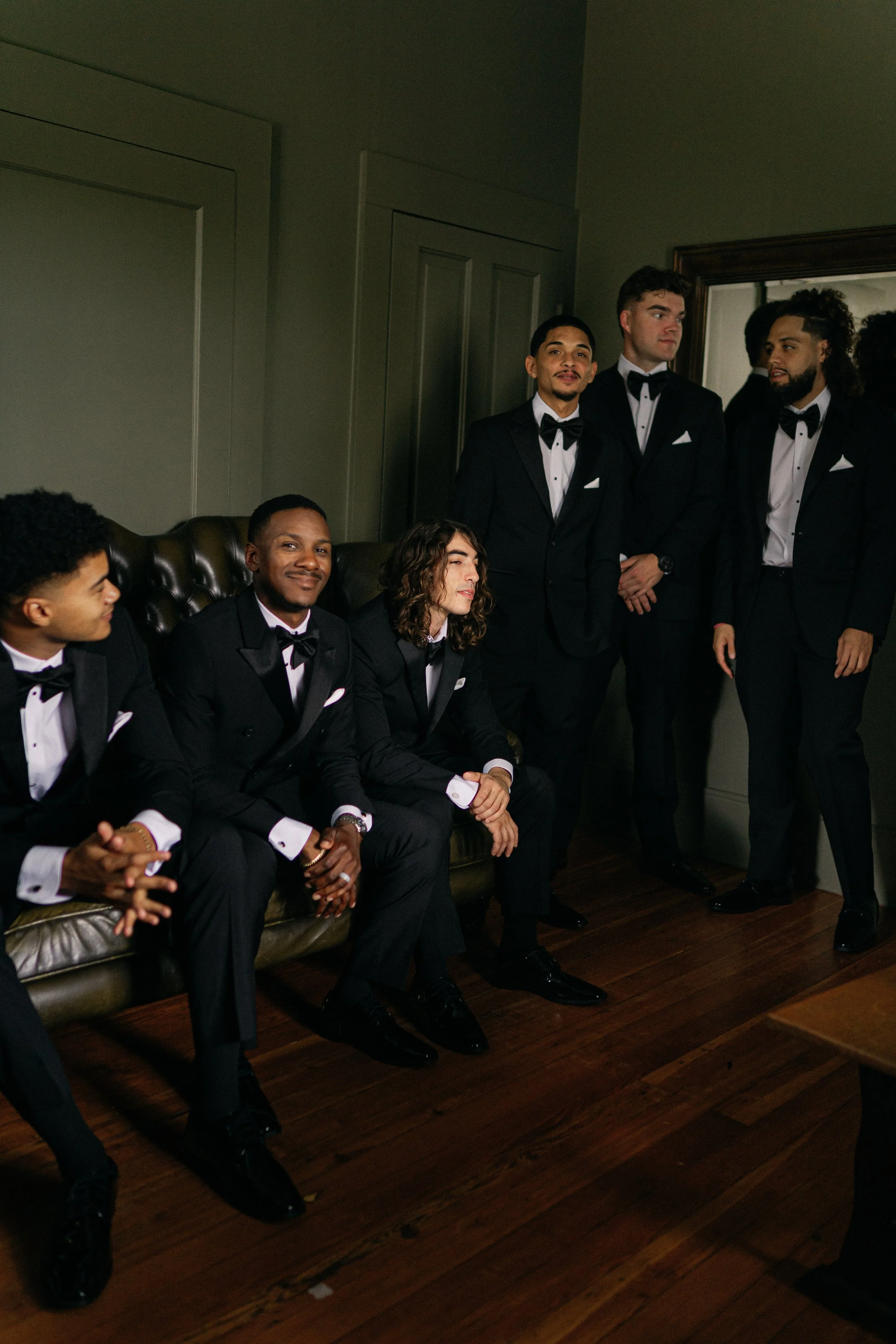 Group of men dressed in black tuxedos with bow ties, sitting and standing in a room with wood flooring and a mirror on the wall. Palm Mansion, Umatilla FL wedding venue. Groomsmen portraits. Luxury wedding. NY photographer