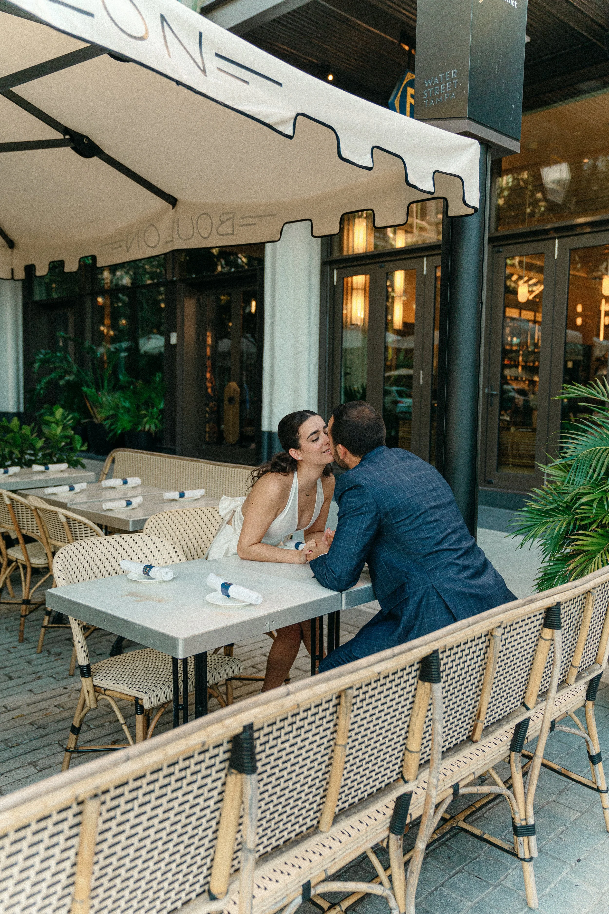 A romantic couple sitting at an outdoor restaurant table, leaning in for a kiss, under a white canopy umbrella with the restaurant's name. Bouilon Brasery Engagement session, Waters St. Tamp FL. NYC couple photographer