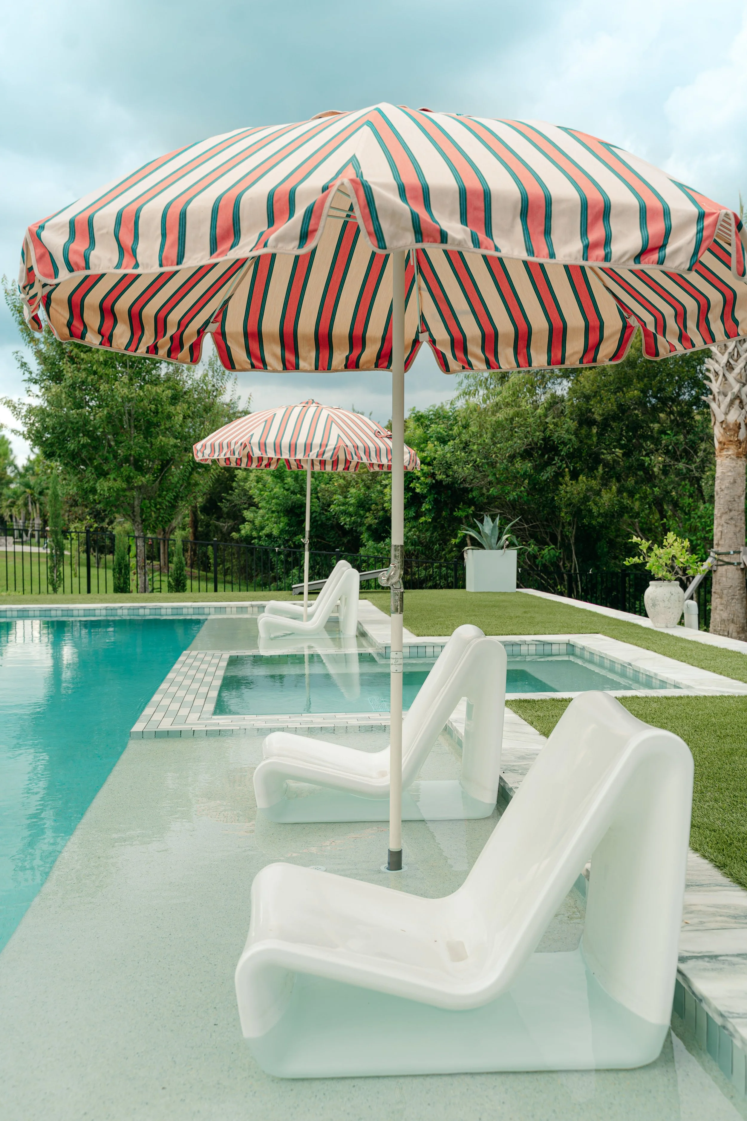 Swimming pool area with white lounge chairs under red, white, and blue striped umbrellas, surrounded by greenery and trees.