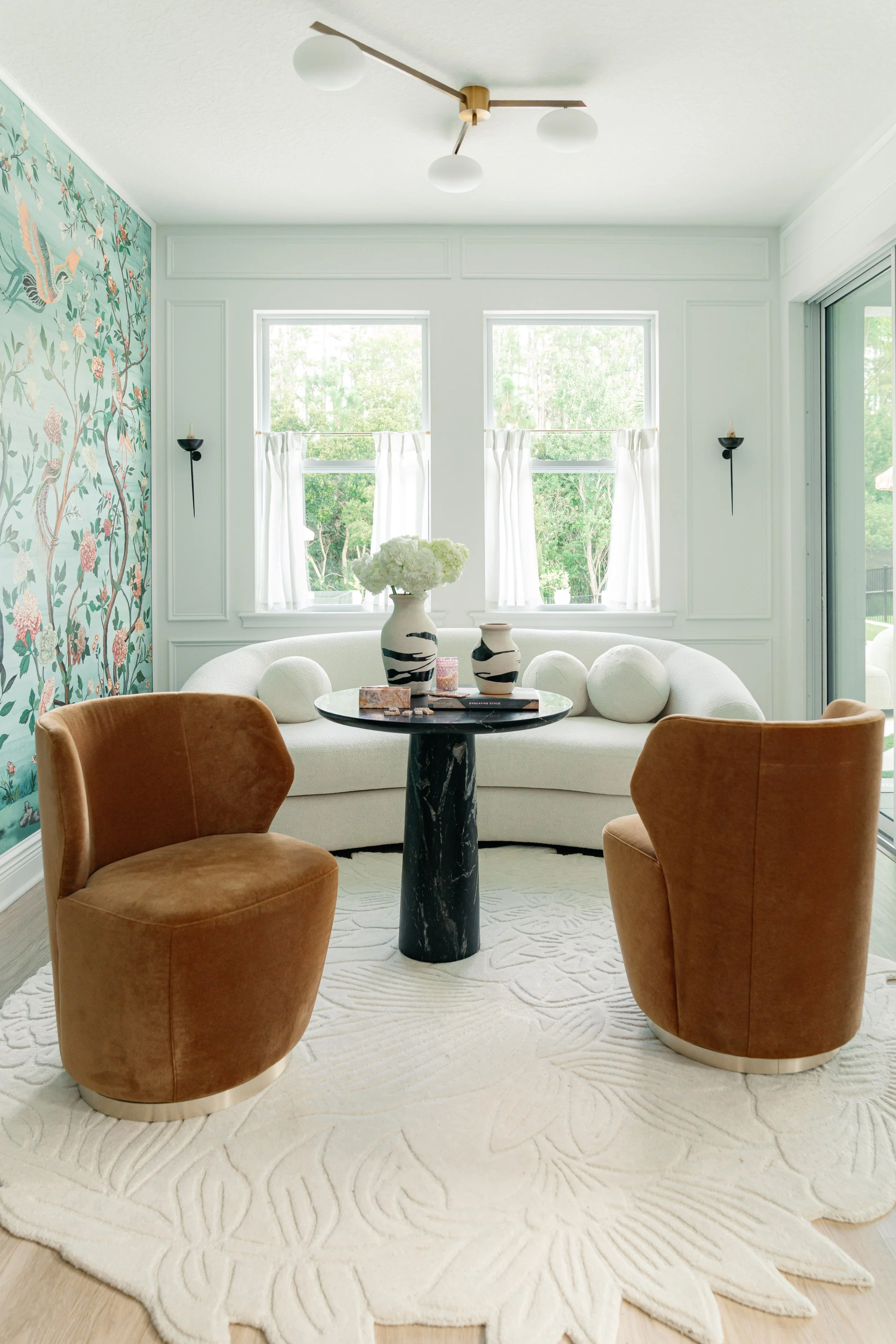Living room with two brown velvet chairs, a white curved sofa, a round black marble table with decorative vases and flowers, floral wallpaper wall, white curtains, and large windows showing green trees outside.