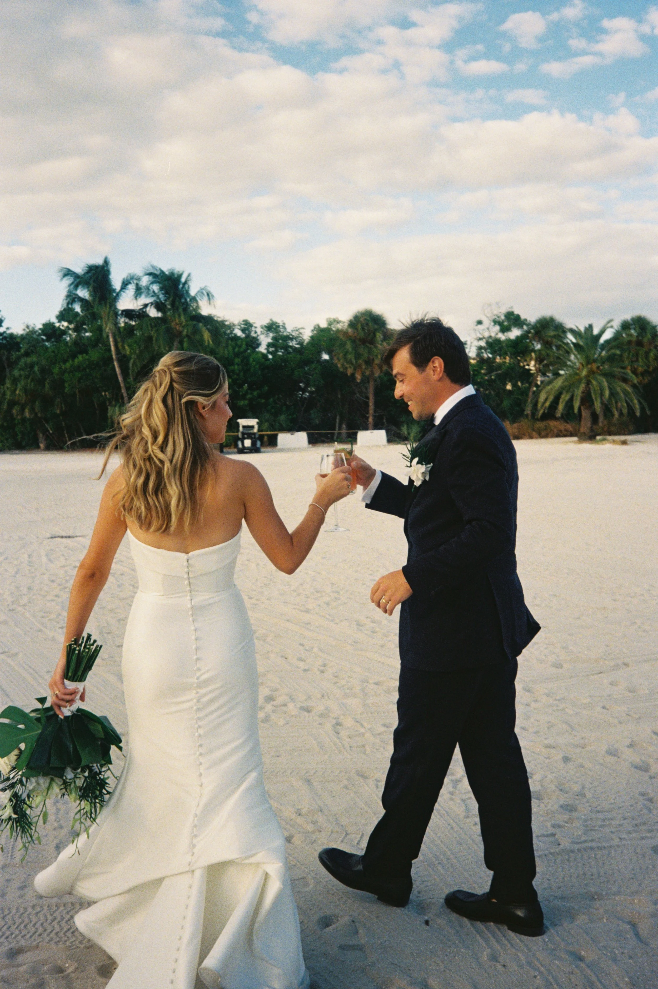 A bride and groom in wedding attire toasting with champagne on a beach with palm trees and a partly cloudy sky. Longboat Key Club venue.  Florida coastal wedding portraits. Film photography