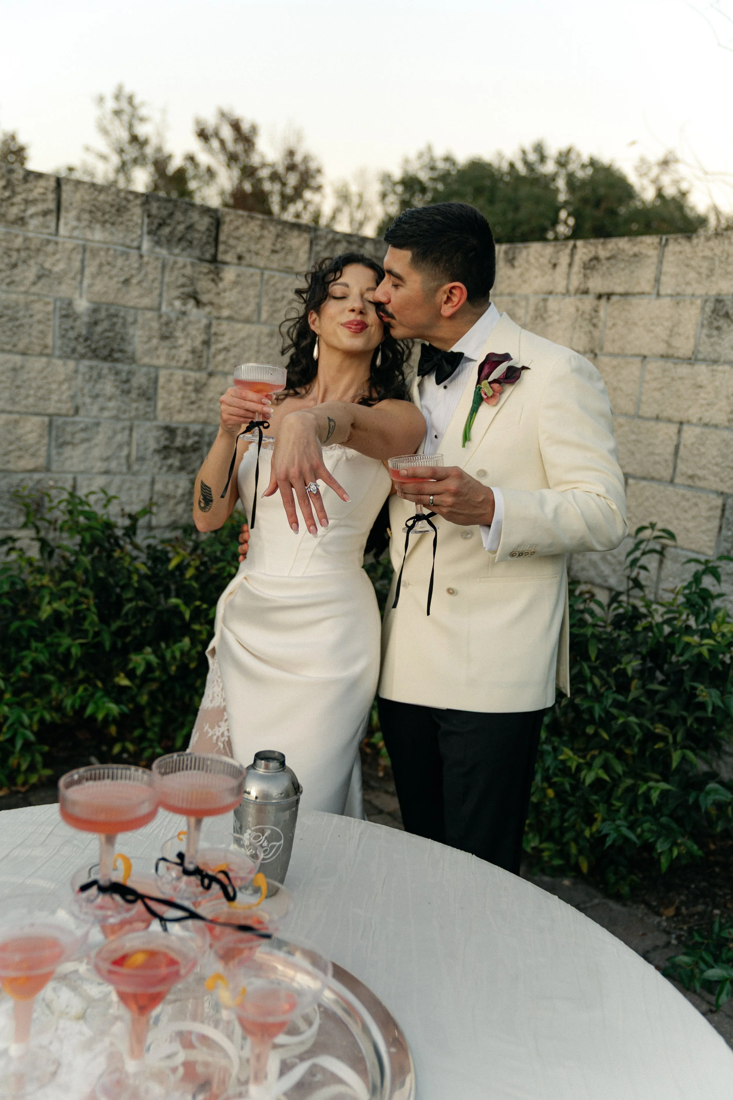 Couple at their wedding reception, with the bride showing her engagement ring and the groom kissing her cheek. They are dressed in formal attire, holding drinks, with a table of cocktails in the foreground. The Secret Garden, Paradise Spring venue.