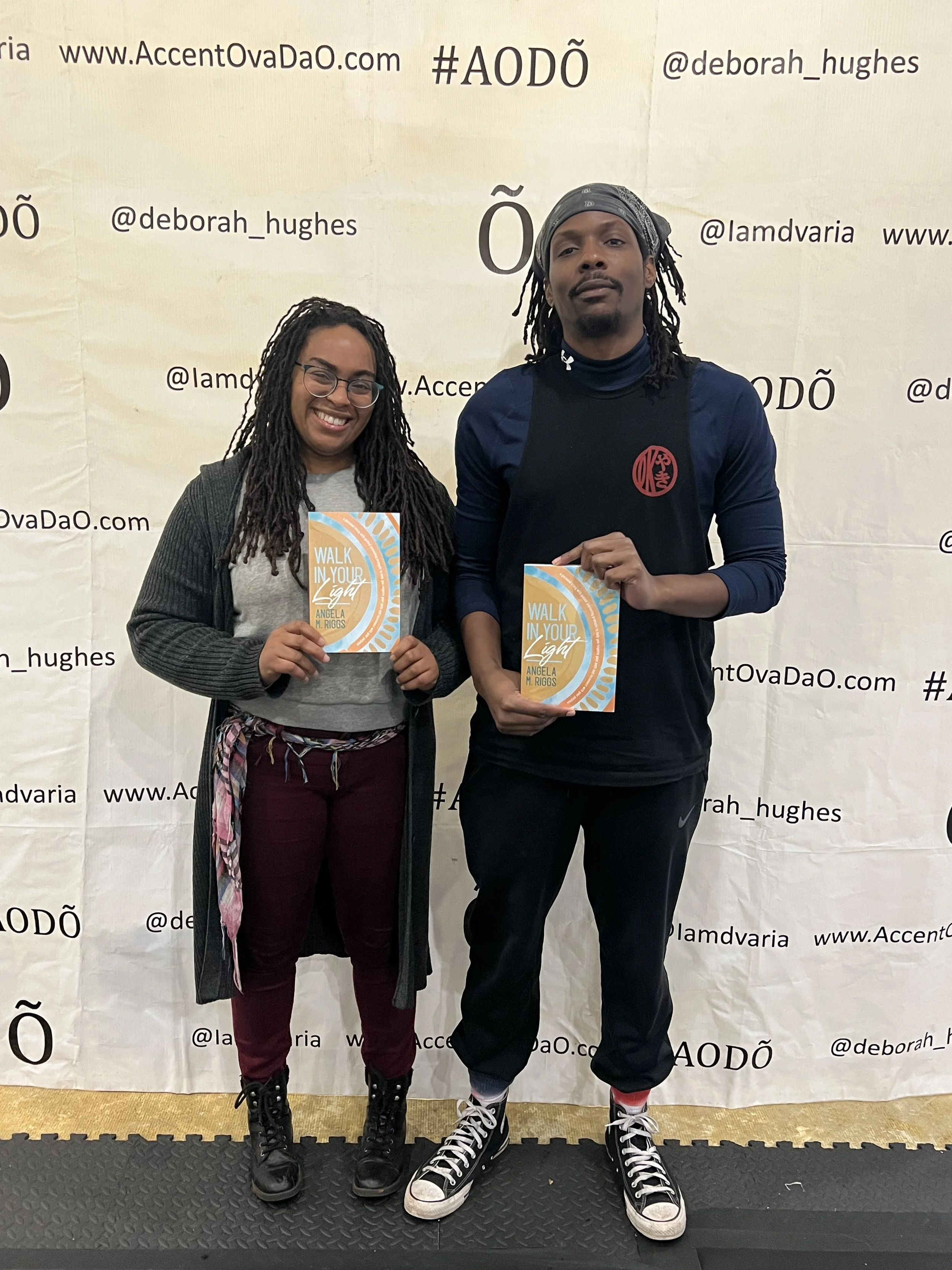 Two people standing in front of a backdrop, each holding a book titled 'Walk in Your Light' by Angela M. Riggs. The person on the left is a woman with long dreadlocks, glasses, and wearing a gray shirt, black jumper, burgundy pants, and black boots. The person on the right is a man with dreadlocks, a bandana, wearing a dark long-sleeve shirt, black pants with white stripe details, and black and white sneakers.
