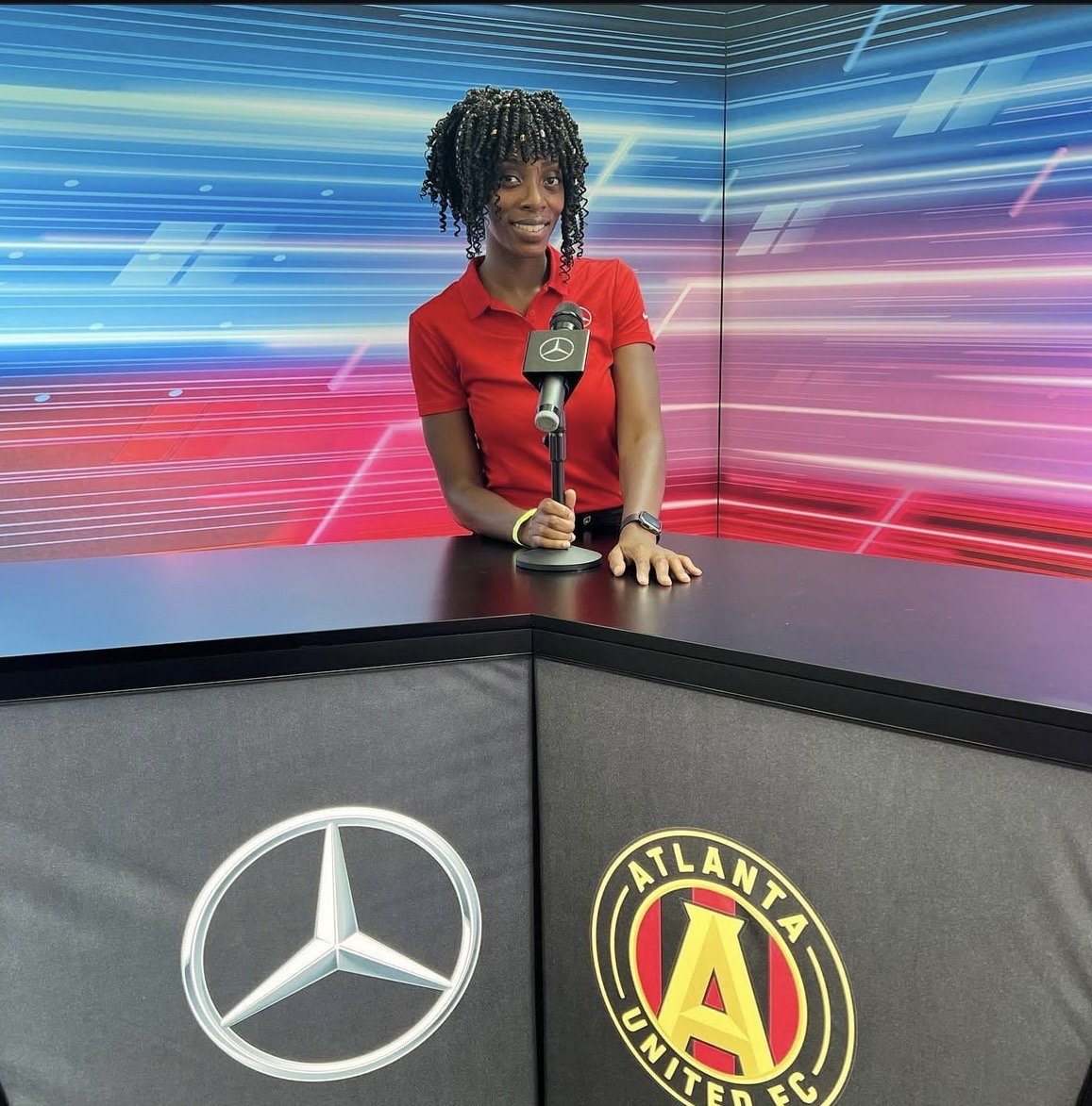 A woman standing behind a desk with a Mercedes-Benz logo and Atlanta United FC logo, holding a microphone, with a vibrant, colorful digital background.