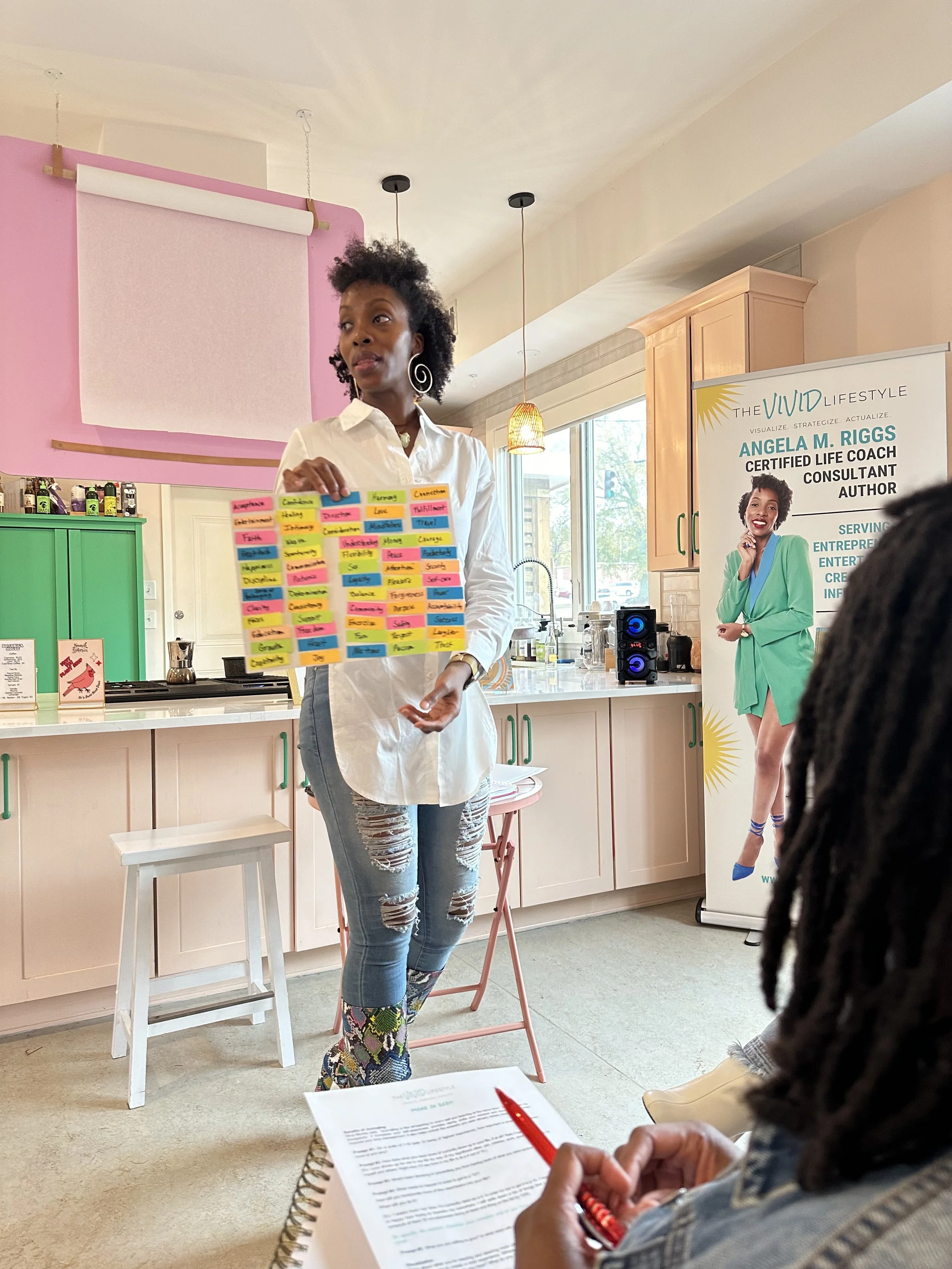 A woman giving a presentation in a kitchen, holding colorful sticky notes with various words. There are people seated, one holding a notebook and pen, and a banner in the background promoting Angela M. Riggs, a certified life coach and author.