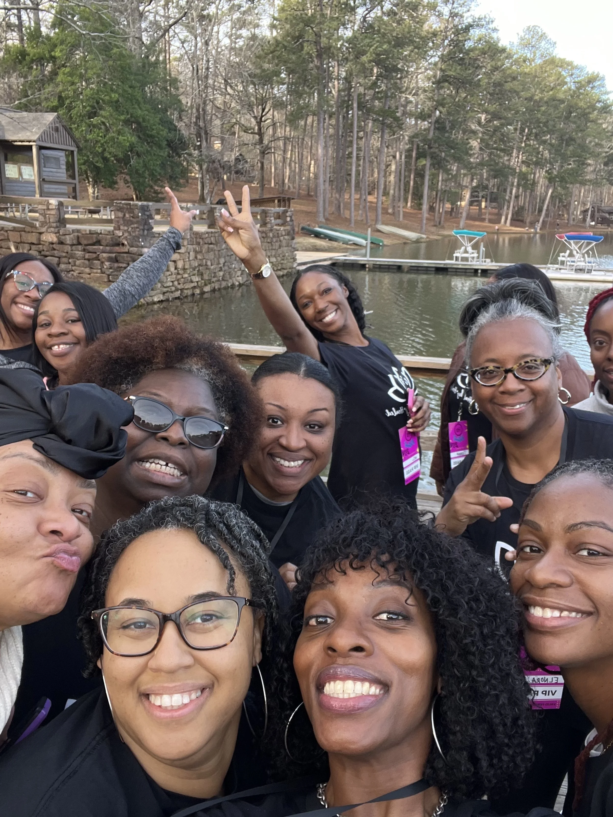 Group of women smiling and taking a selfie by a lake with trees and docks in the background.