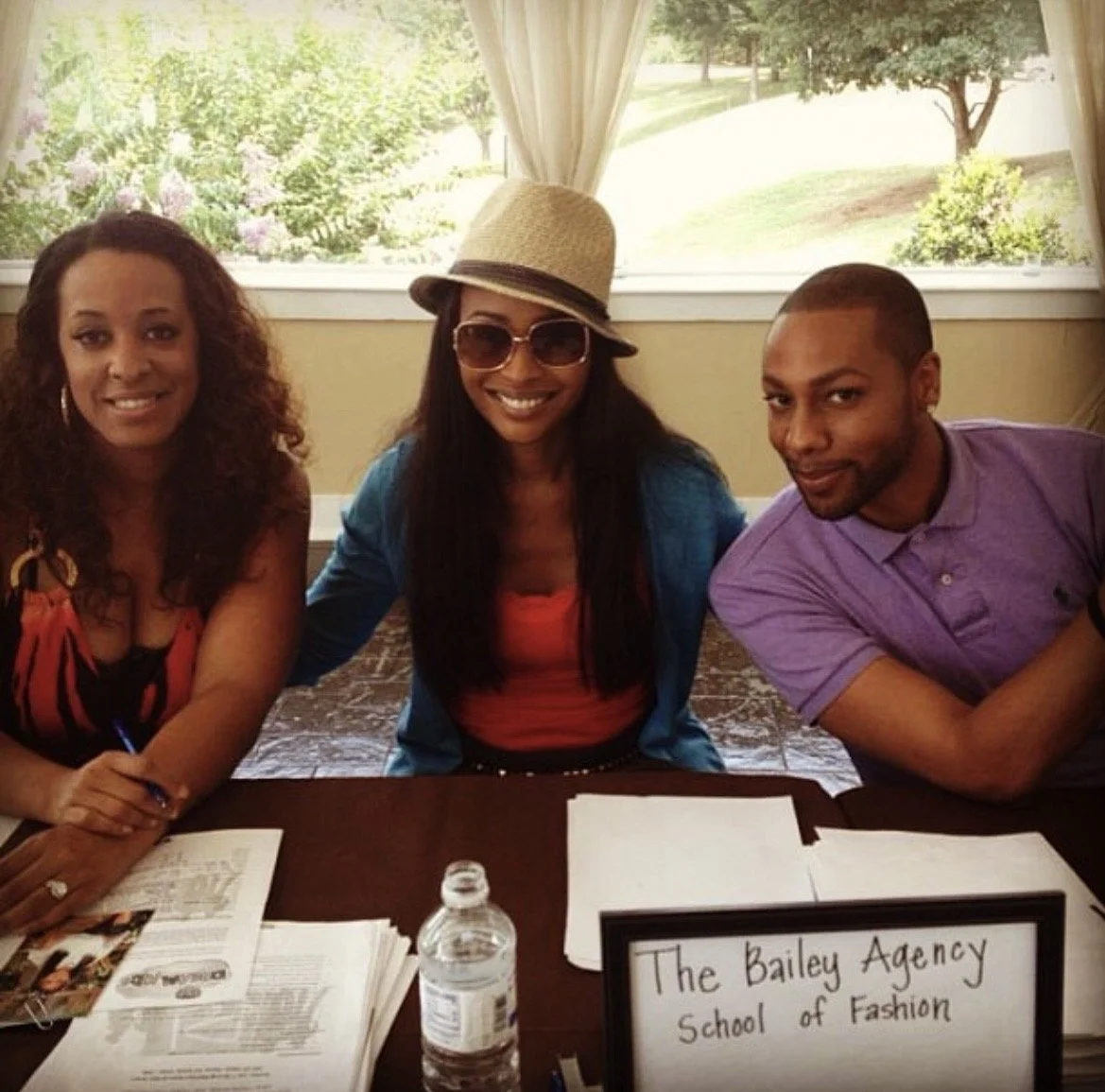 Three smiling individuals, two women and one man, sitting at a table with papers and a water bottle, indoors with a large window showing a garden outside. A sign on the table reads 'The Bailey Agency School of Fashion.'