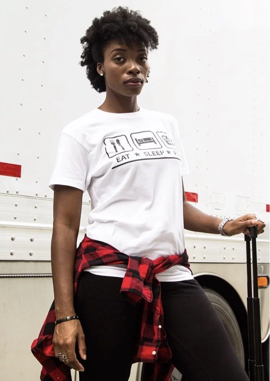 A woman with natural curly hair standing in front of a white truck, wearing a white T-shirt that says "Eat Sleep" with icons of a fork, plate, and a bed, and black pants with a red and black plaid shirt tied around her waist, holding a black suitcase.