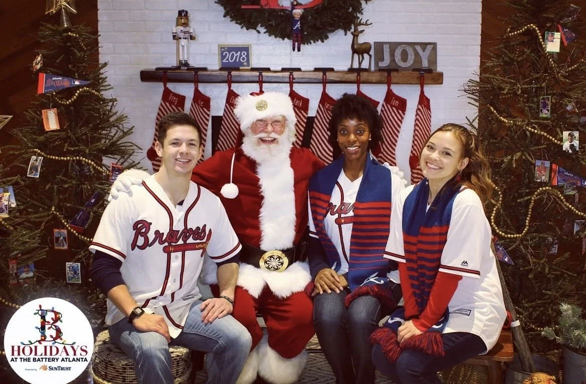 Four people, three young adults and Santa Claus, are sitting together in front of Christmas decorations, including Christmas trees, stockings, and a wreath. They are smiling and dressed in Braves baseball jerseys and holiday attire.