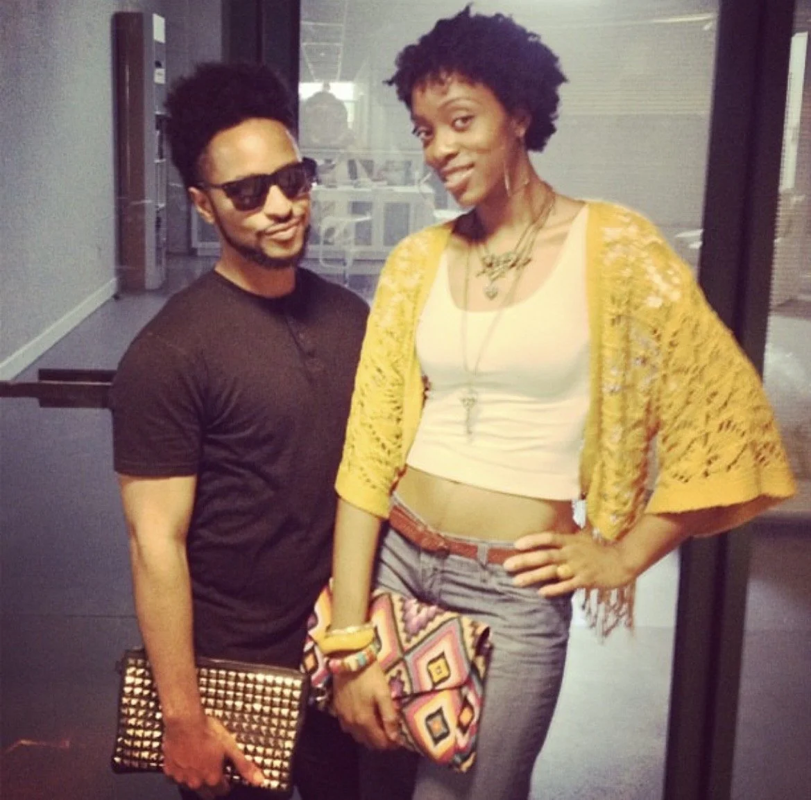 A man and woman standing indoors, smiling at the camera, with the woman slightly taller and have curly short hair. The man is wearing a black shirt, sunglasses, and holding a brown clutch, while the woman is wearing a white top, yellow cardigan, plaid shorts, and carrying a colorful clutch. The woman has multiple necklaces and bracelets.