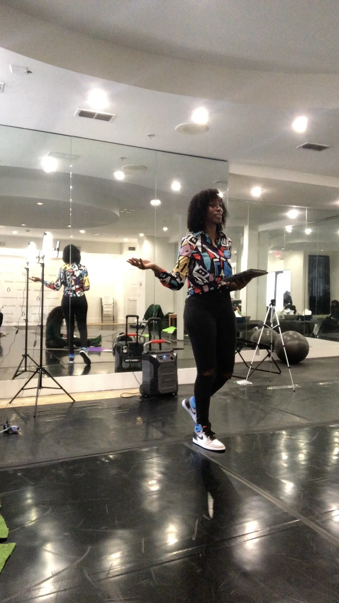 A woman standing on a stage with a mirror behind her, holding a tablet, with studio lighting and exercise equipment in the background.