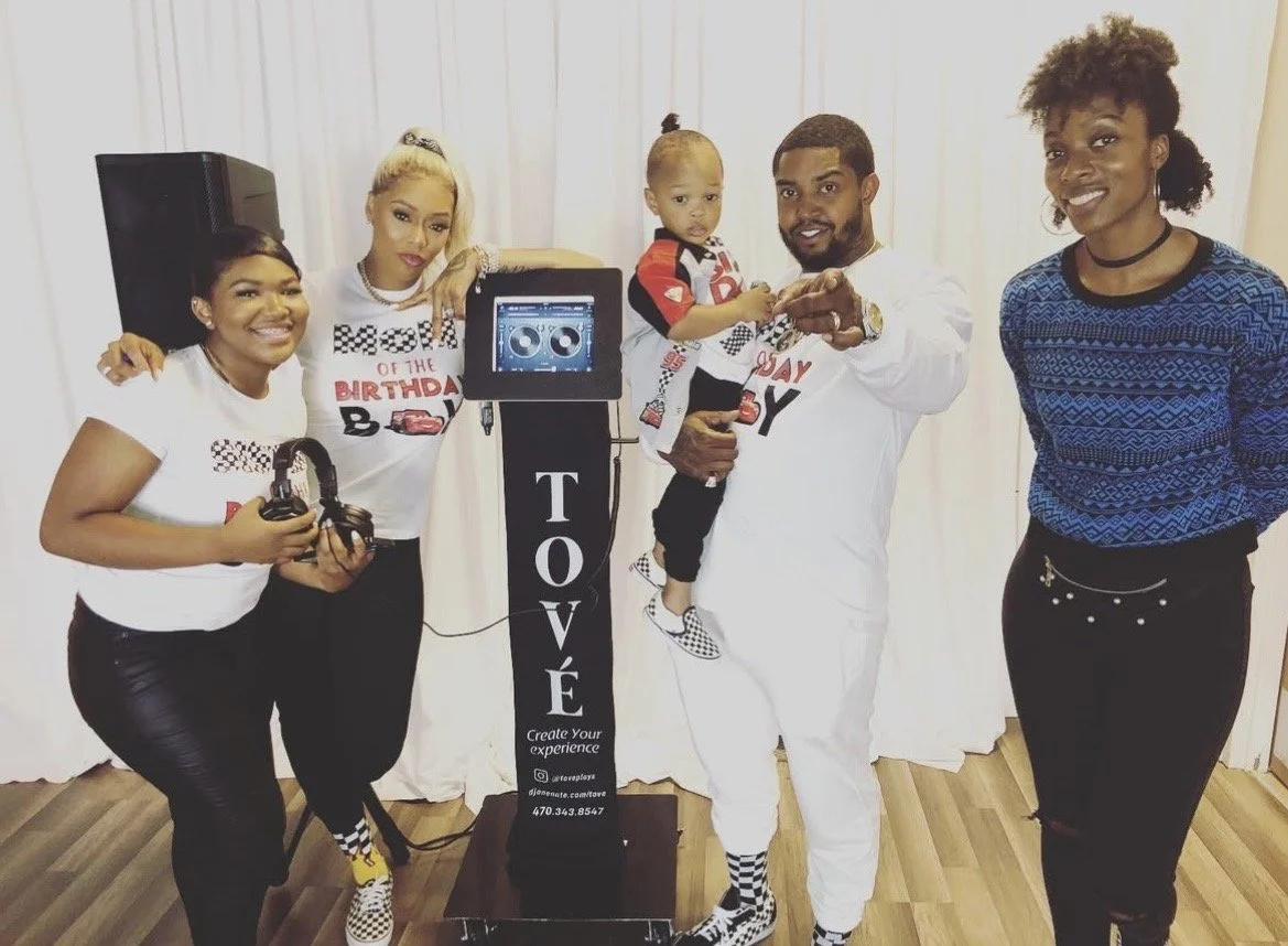 Group of five people celebrating a birthday, with a man holding a young child, standing next to a birthday cake-shaped photo booth labeled 'TOVÉ'.