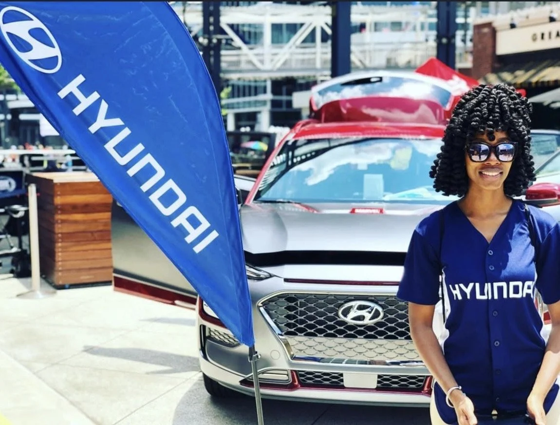 A woman with curly hair and sunglasses standing in front of a silver Hyundai car at an outdoor event, with a blue Hyundai flag nearby.