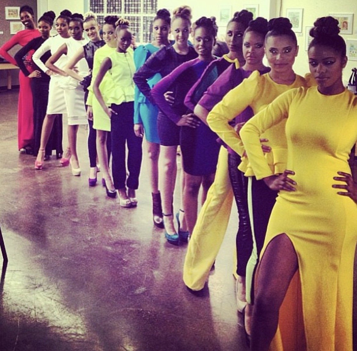 A group of women lined up in a room, posing with hands on hips. They are wearing colorful dresses and high heels, with some in traditional or formal attire. The background shows framed pictures on the wall.