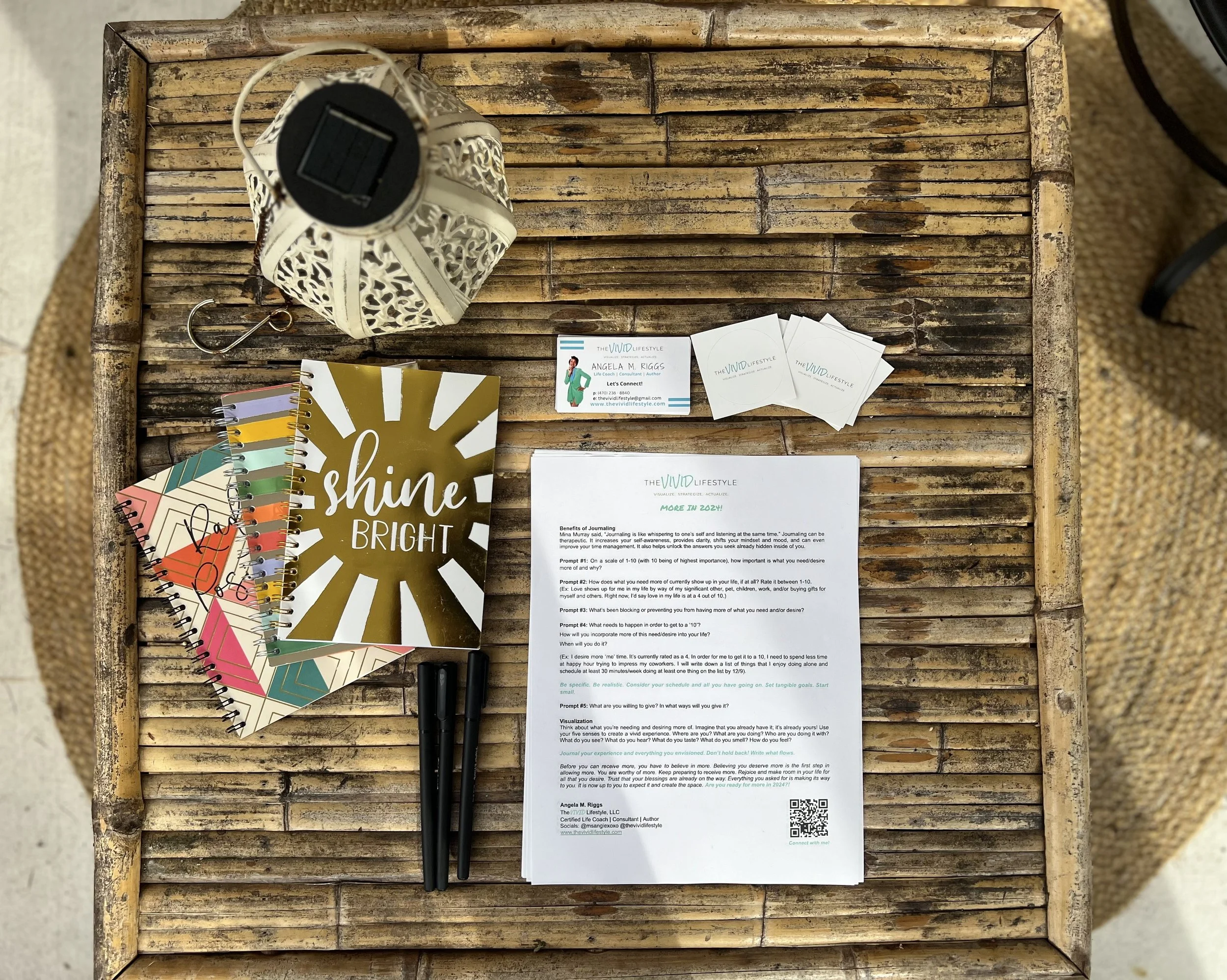A wooden table with various office items including colorful notebooks, business cards, a sheet of paper with text, a couple of black pens, a decorative white lantern, and small packets labeled 'The VIVID Lifestyle'.