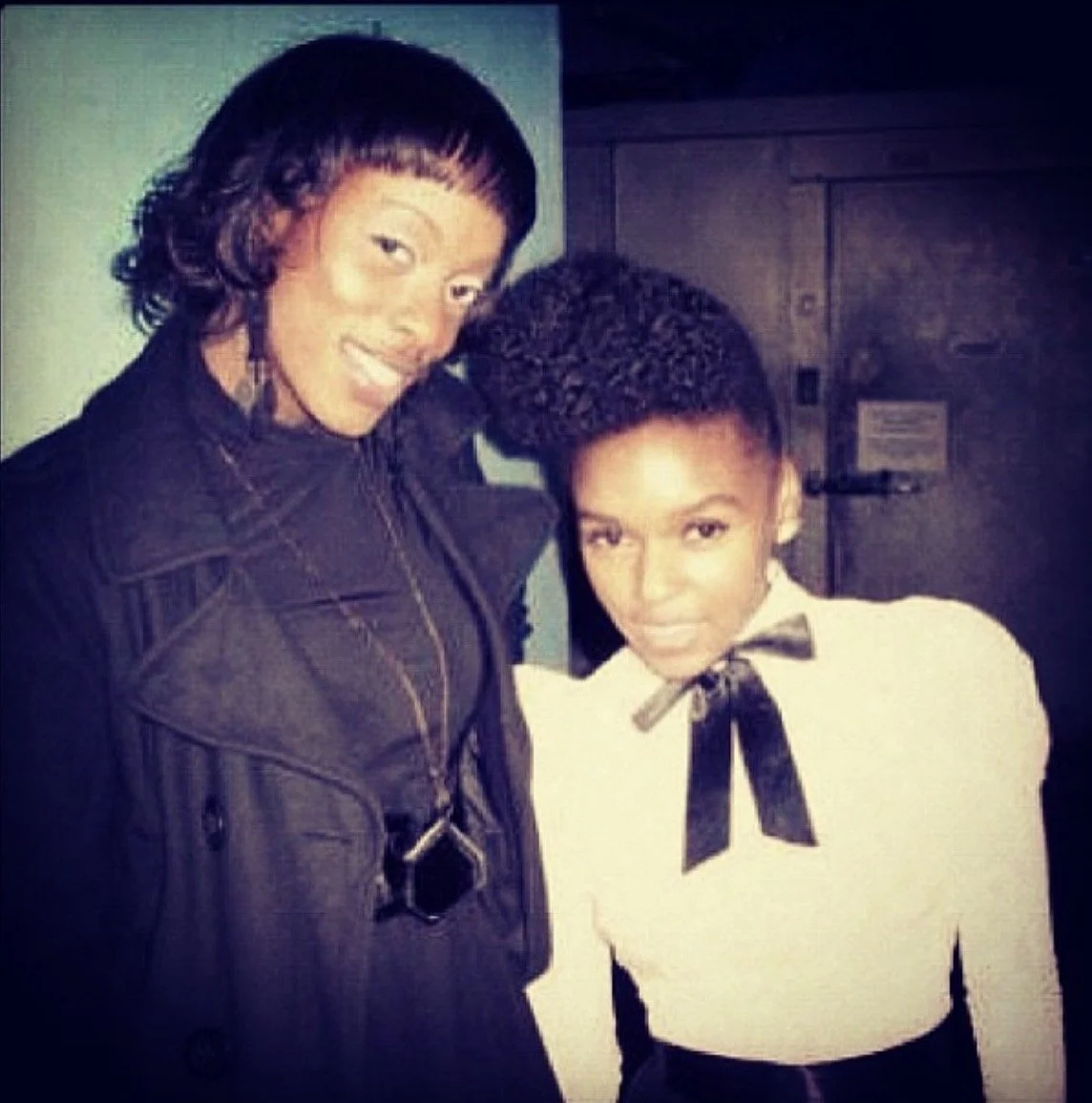 Two women with styled hair standing together, smiling and posing for the photo indoors.