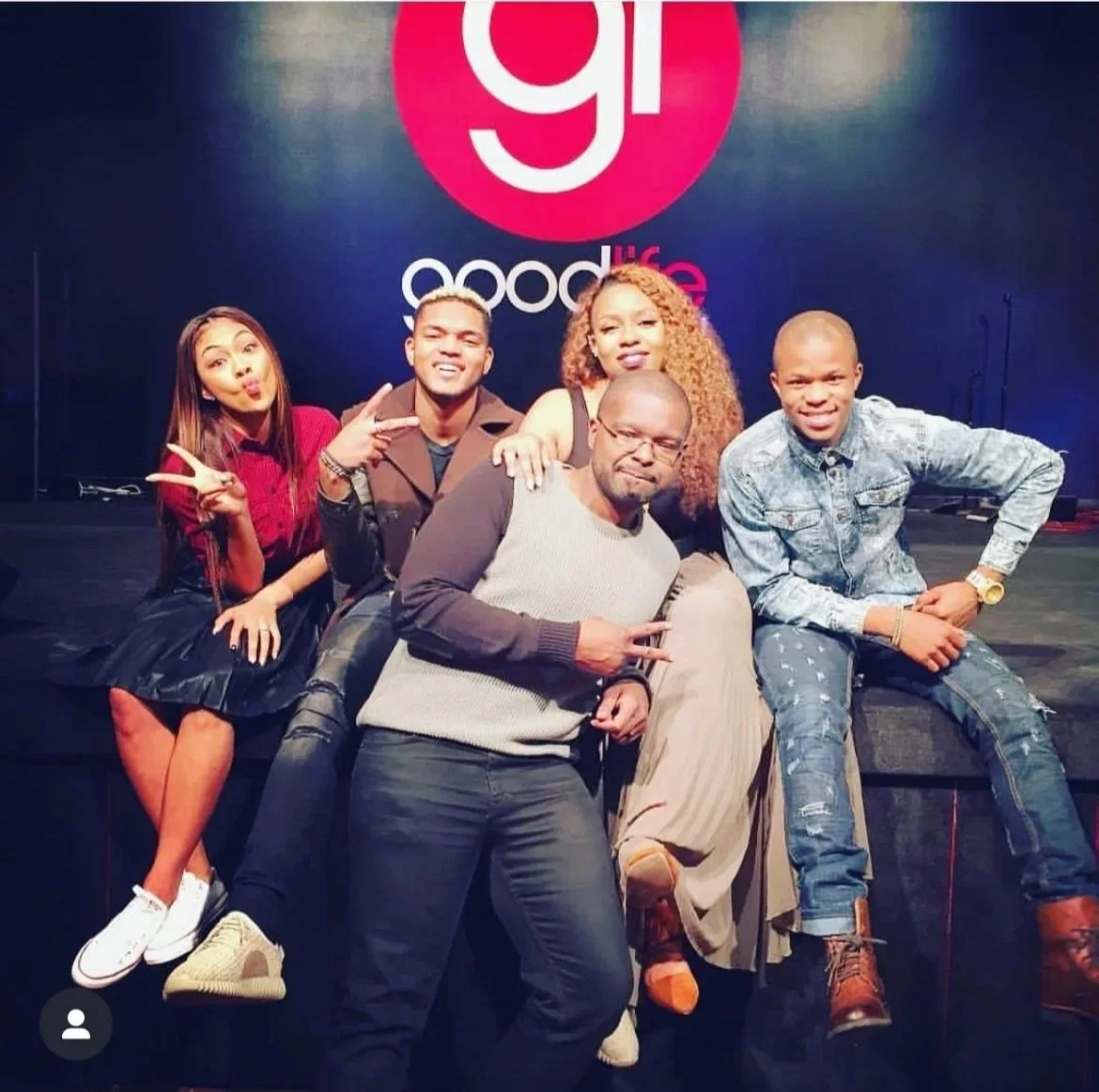 Five people posing on a stage in front of a large logo that says '91' and 'Goodlife'. One woman on the left is making a peace sign, and the others are smiling or making hand gestures.
