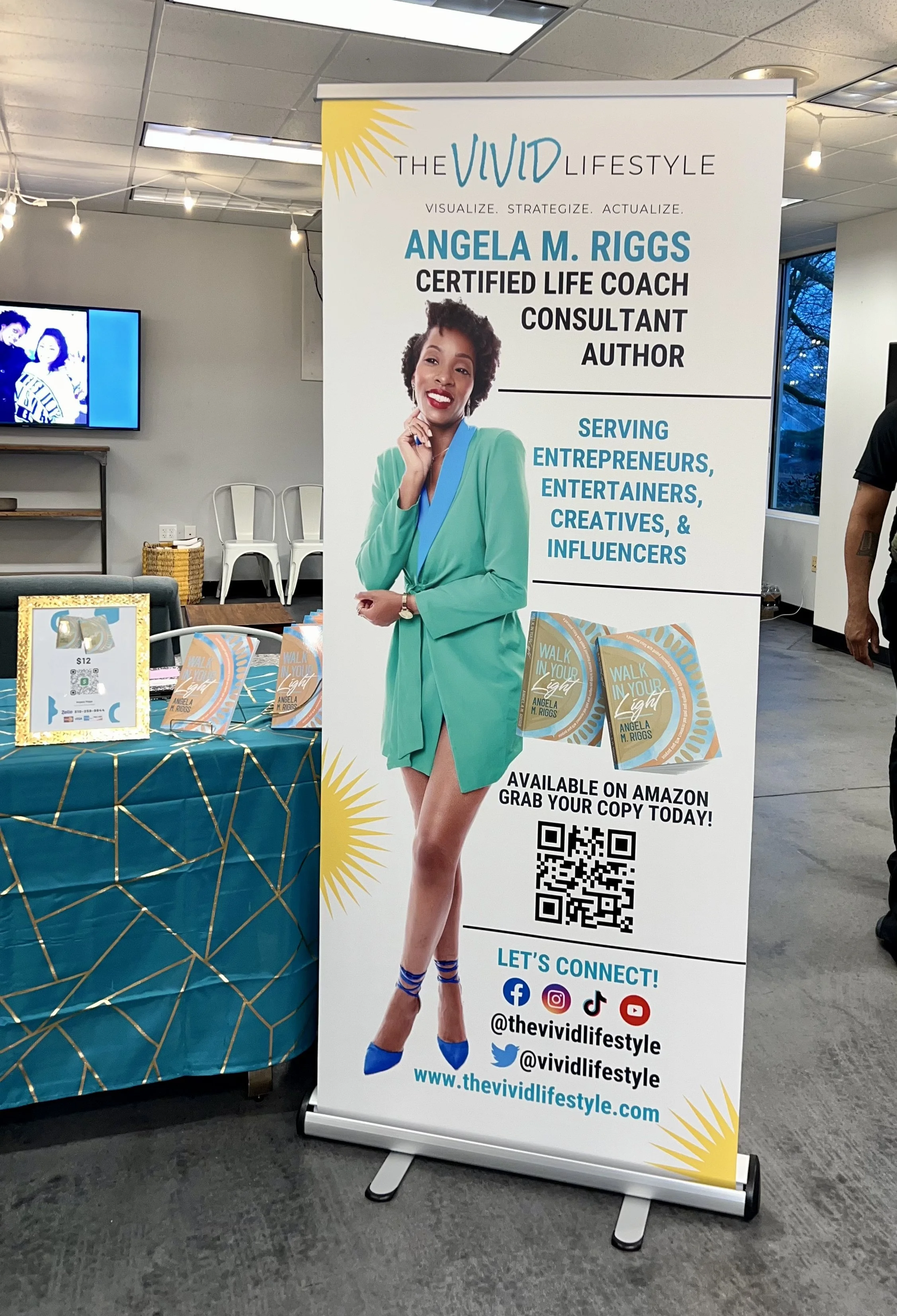 Promotional display for Angela M. Riggs, a certified life coach, author, and consultant, featuring her photo and book cover for 'Walk in Your Light,' with contact and social media information, and a QR code for purchasing on Amazon.