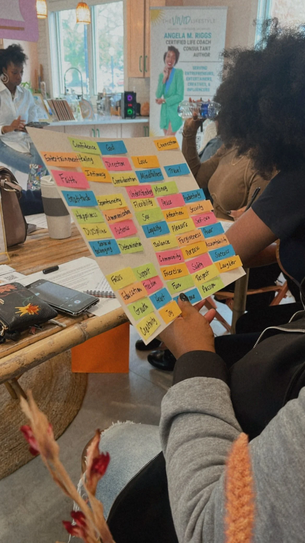 A woman is holding a colorful poster with words on sticky notes, including 'Confidence,' 'God,' 'Connection,' 'Love,' 'Mindfulness,' 'Truth,' 'Happiness,' 'Discipline,' 'Balance,' 'Community,' and 'Success.' The setting appears to be a workshop or seminar with several people seated around a table in a cozy, well-lit room.