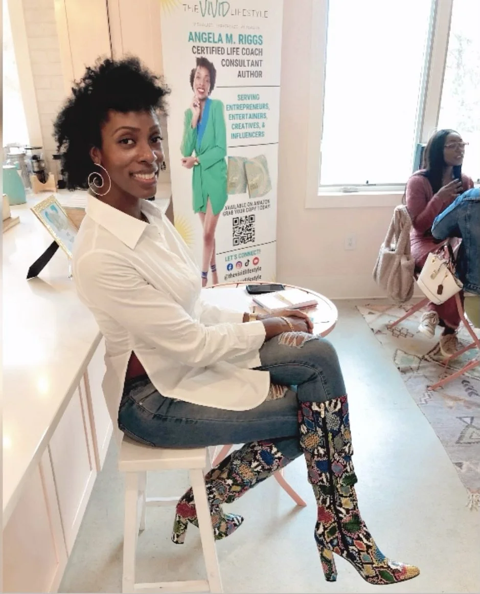 A woman sitting on a white stool wearing a white shirt, jeans, and colorful patterned high-heeled boots in a well-lit room with a window. Behind her is a banner featuring Angela M. Riggs, a certified life coach, author, and consultant. Other people in the background are seated, and the room has a light, modern decor.