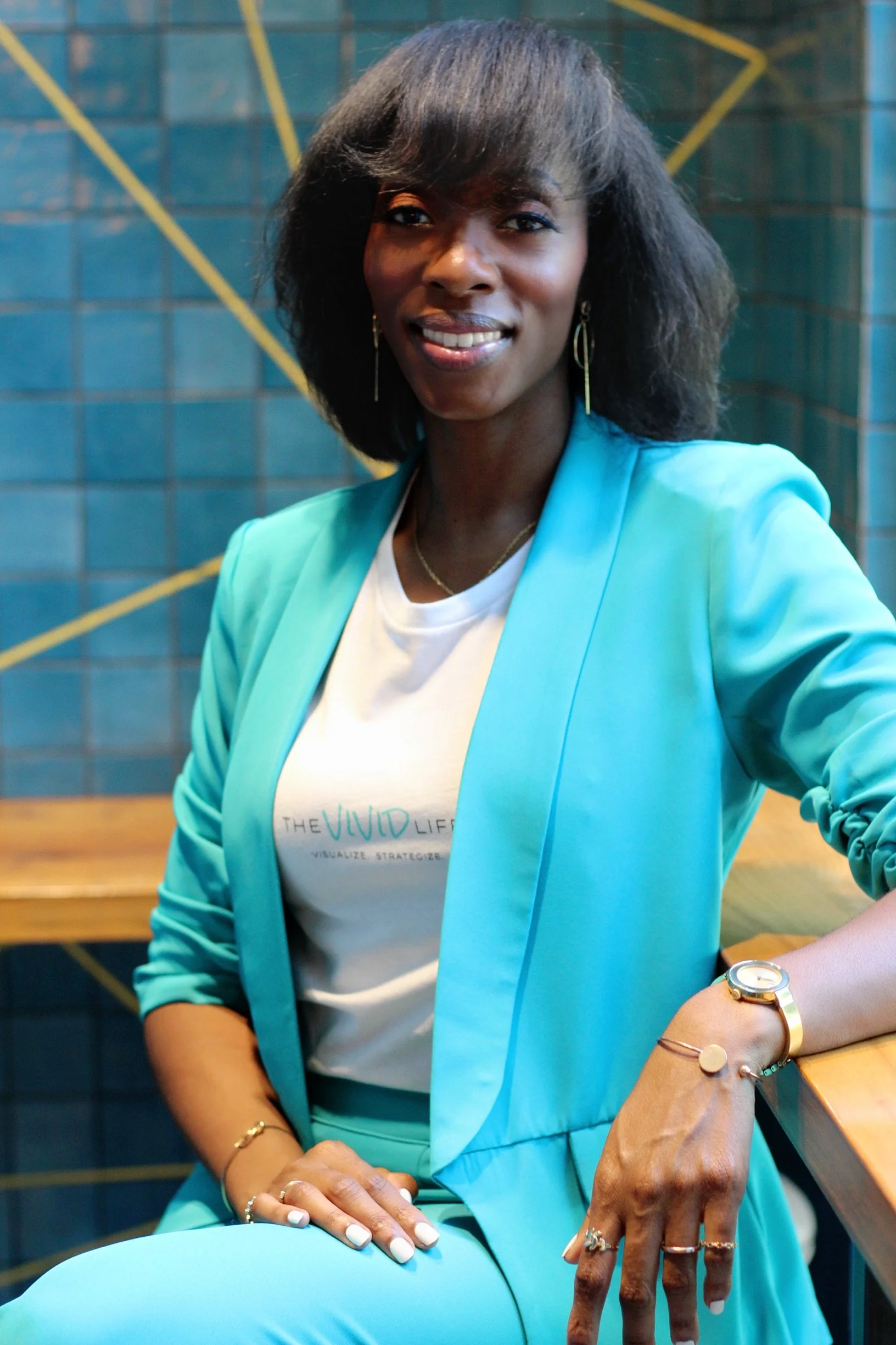 A woman with dark skin and medium-length black hair, wearing a light blue blazer and matching pants, sitting at a wooden table in front of a blue tiled wall with yellow lines.