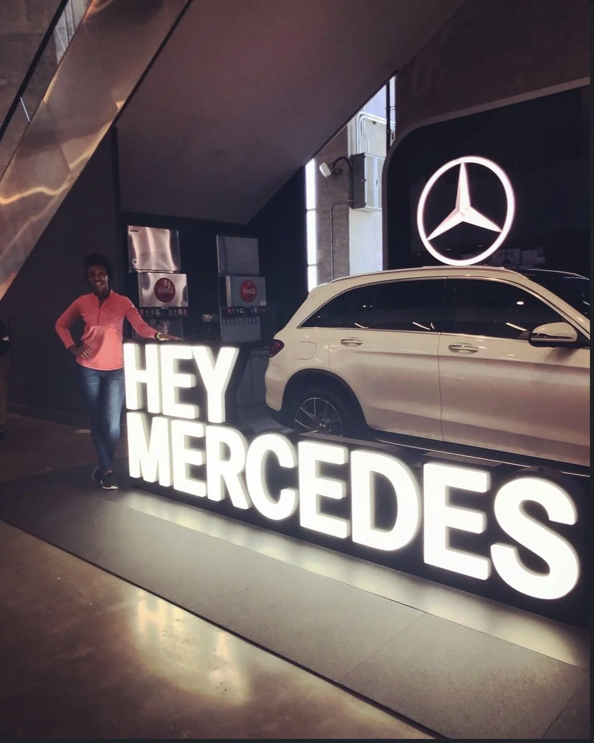 A woman standing next to a large illuminated sign that reads 'HEY MERCEDES' in a showroom with a Mercedes-Benz vehicle and a Mercedes logo display in the background.