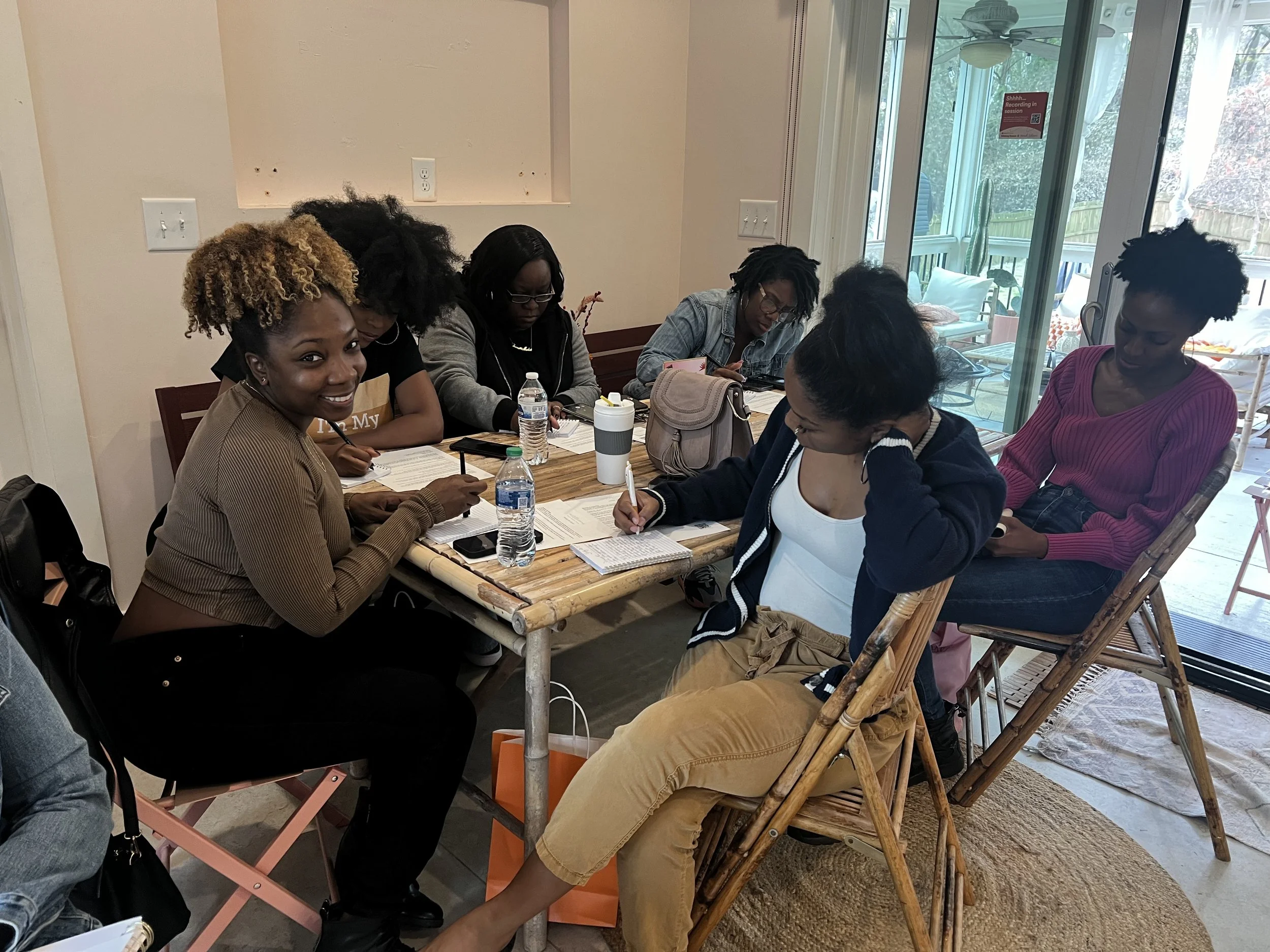 A group of seven women sitting around a wooden table in a bright room, some writing or looking at papers, with bottles of water and a coffee tumbler on the table. They are engaging in a meeting or discussion. There is a sliding glass door in the background leading to an outdoor patio.