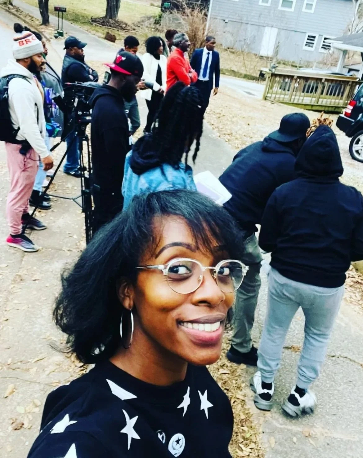 A woman smiling at the camera while taking a selfie outdoors with a group of people gathered behind her, some are looking at a display or event in a residential area during the daytime.