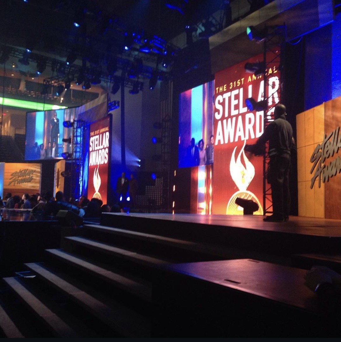 A stage decorated for the Stellar Awards with large screens displaying the event's logo, a flame with a star, and people on stage preparing for the event.