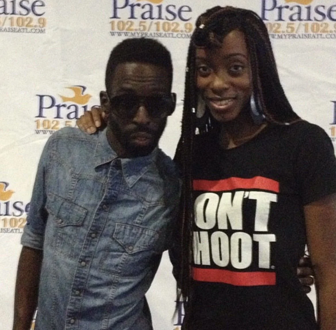 Two people standing in front of a Praise 102.5/102.9 radio station backdrop, posing for the photo. The man is wearing sunglasses and a denim shirt, and the woman is wearing a black T-shirt with bold white and red text.
