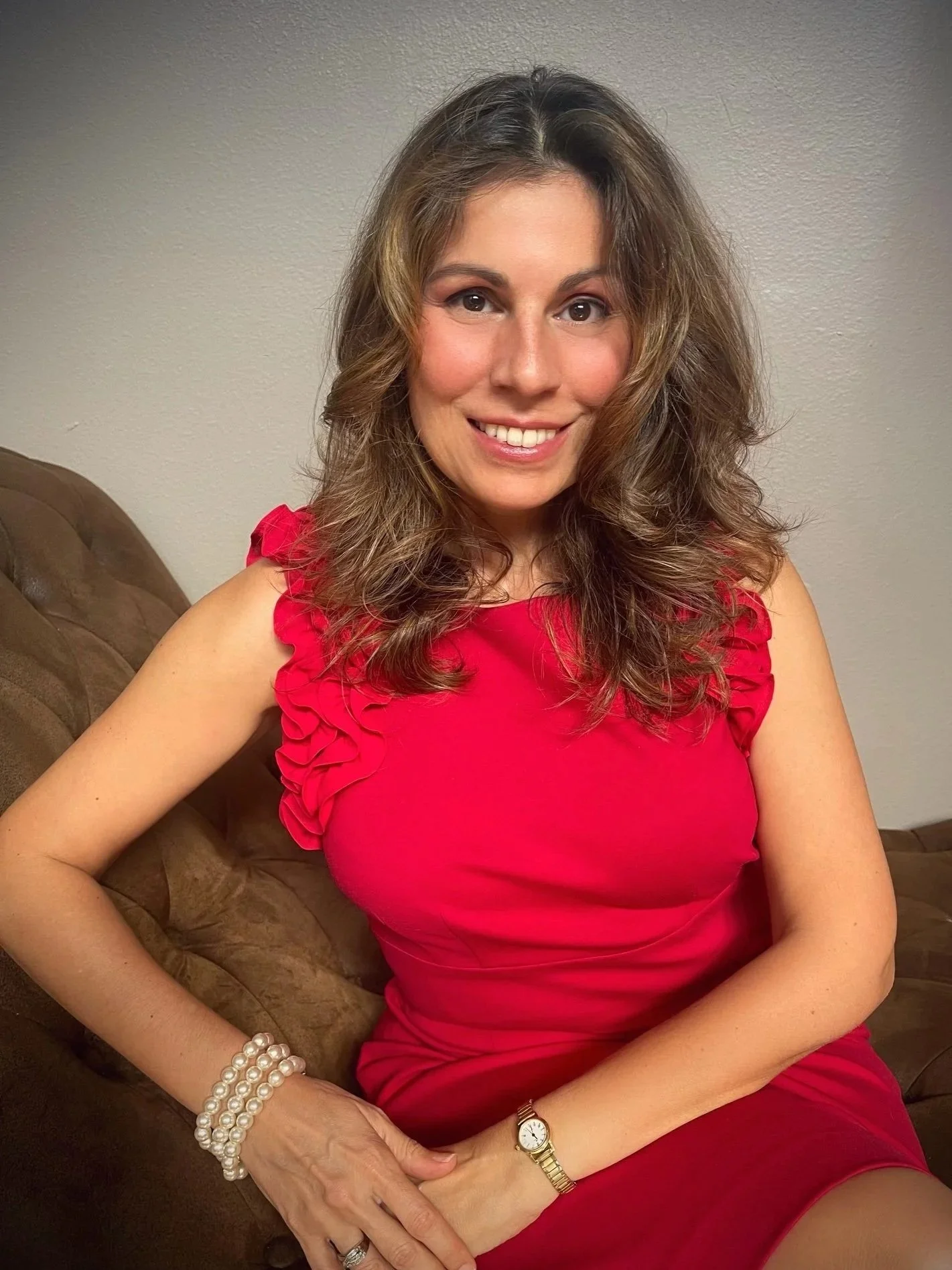 A woman with brown hair and a warm smile is sitting on a brown couch, wearing a red dress with ruffled sleeves. She has pearl bracelets on her wrist, a gold watch, and a ring on her finger. The background is a plain light-colored wall.