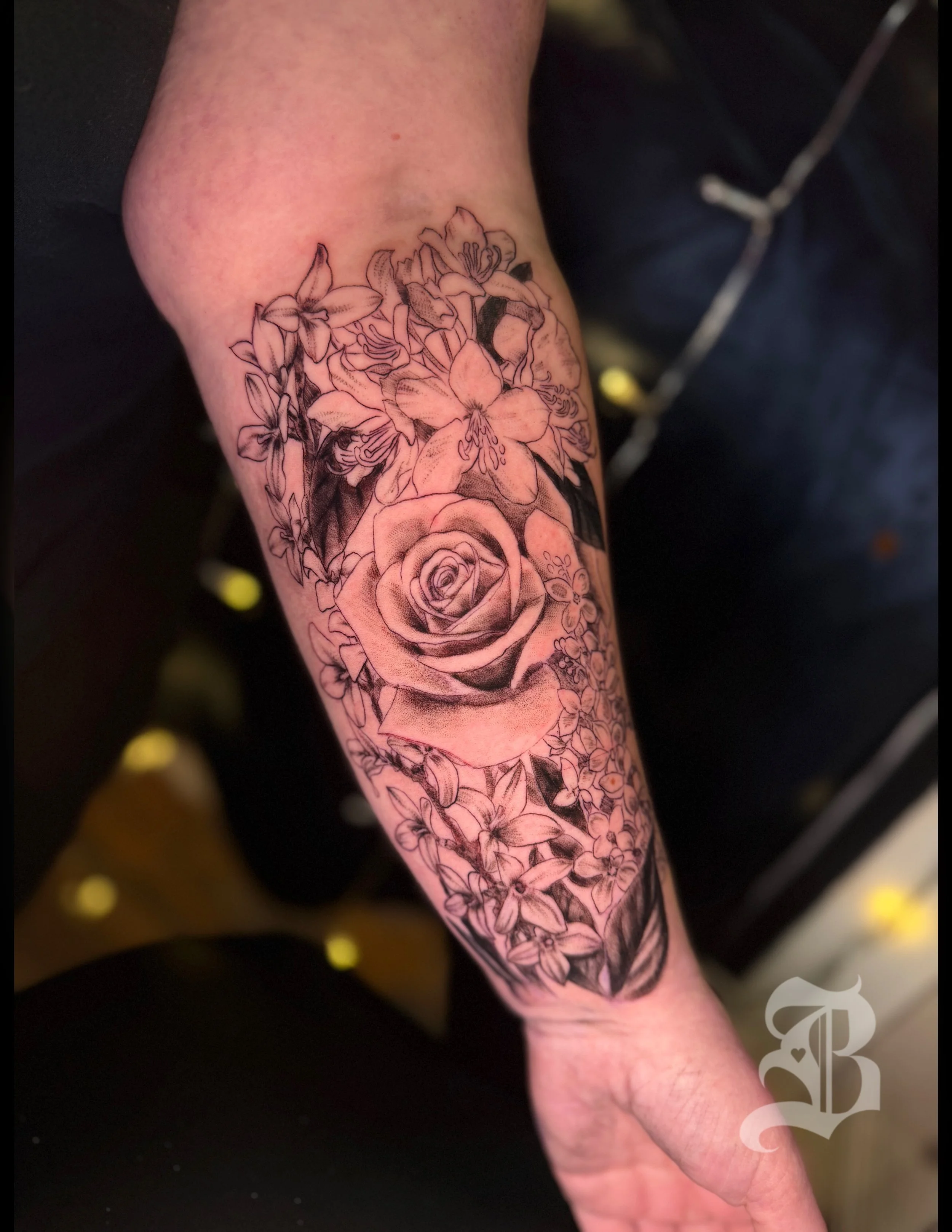 Black and gray floral tattoo featuring a rose surrounded by lilies and other flowers on a person's forearm.