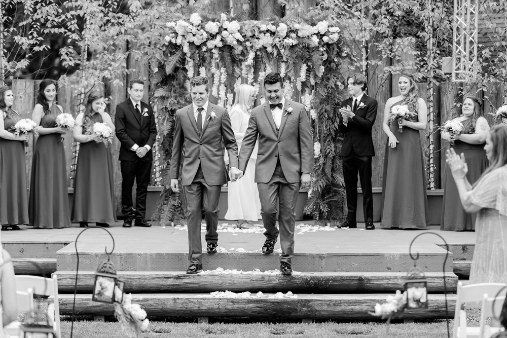 Black and white photo of two grooms recessing down the aisle holding hands after getting married. 