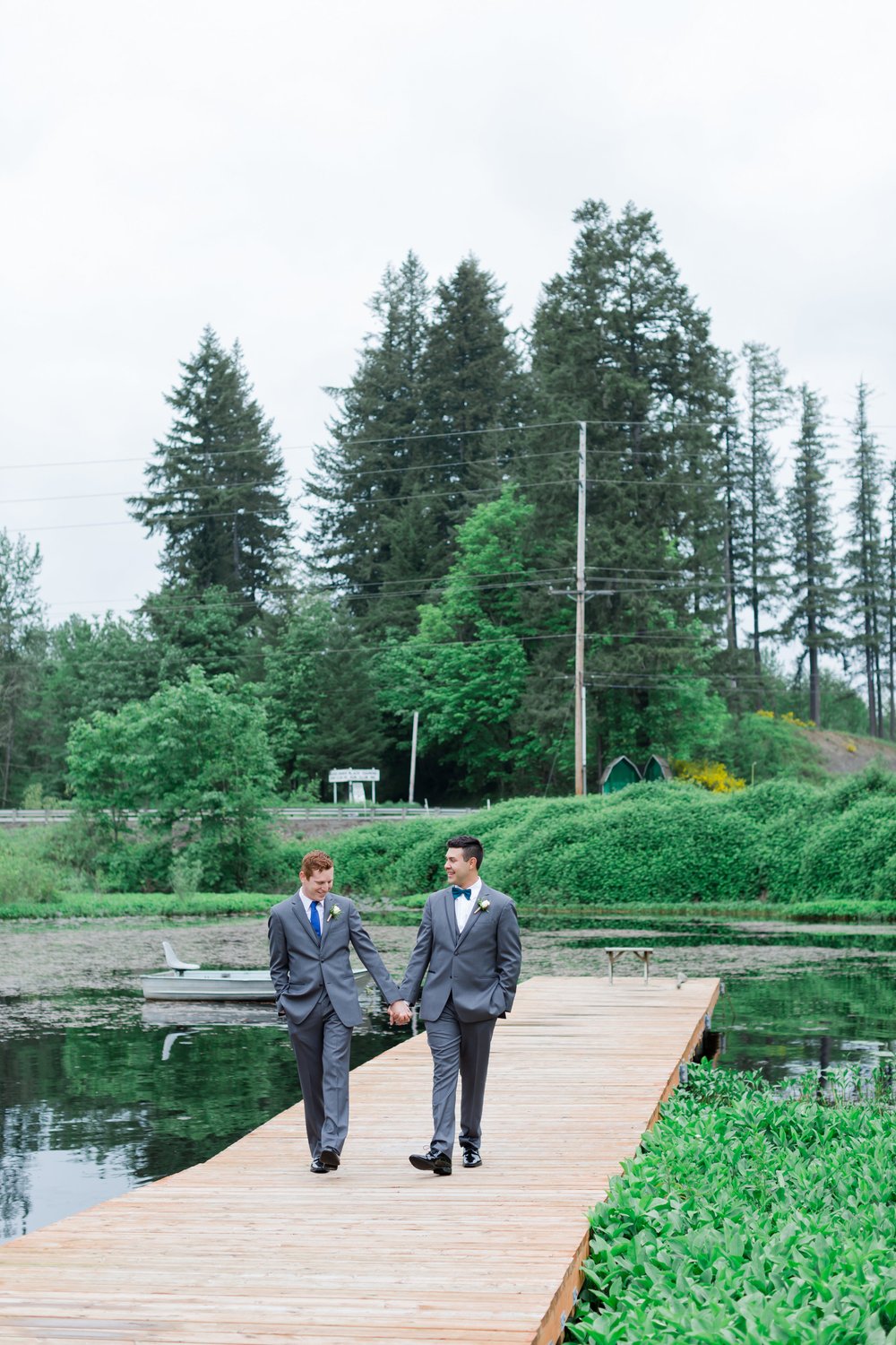  Two grooms wearing gray suits hold hands on a dock on a lake. 